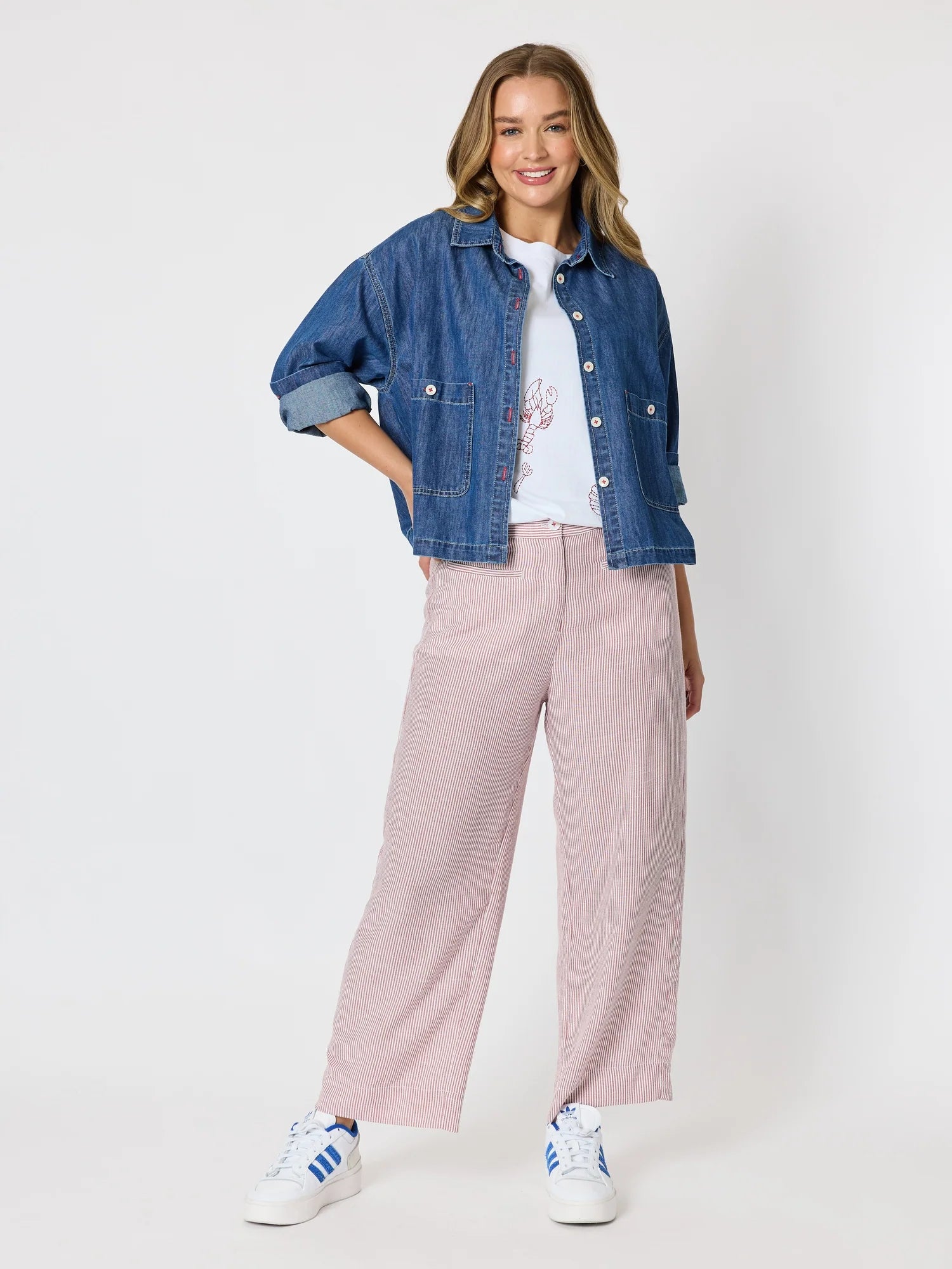 Image of A woman modeling a blue cropped denim jacket over a white graphic t-shirt, paired with Threadz light pink wide-leg trousers and white sneakers with blue stripes, set against a plain light gray studio background.