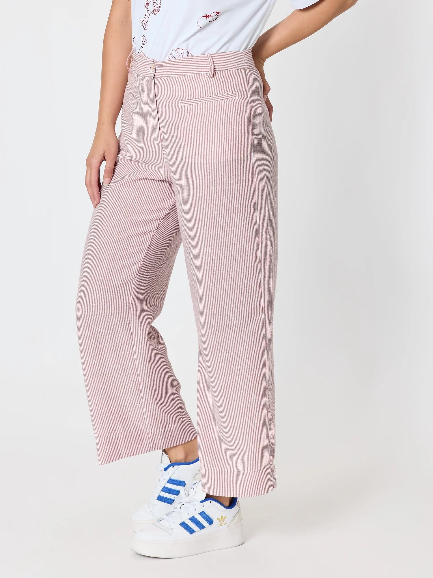Image of A woman wearing Threadz light pink wide-leg cropped trousers with a subtle vertical textured stripe pattern, styled with a white graphic t-shirt and white sneakers with blue stripes, against a plain white studio background.