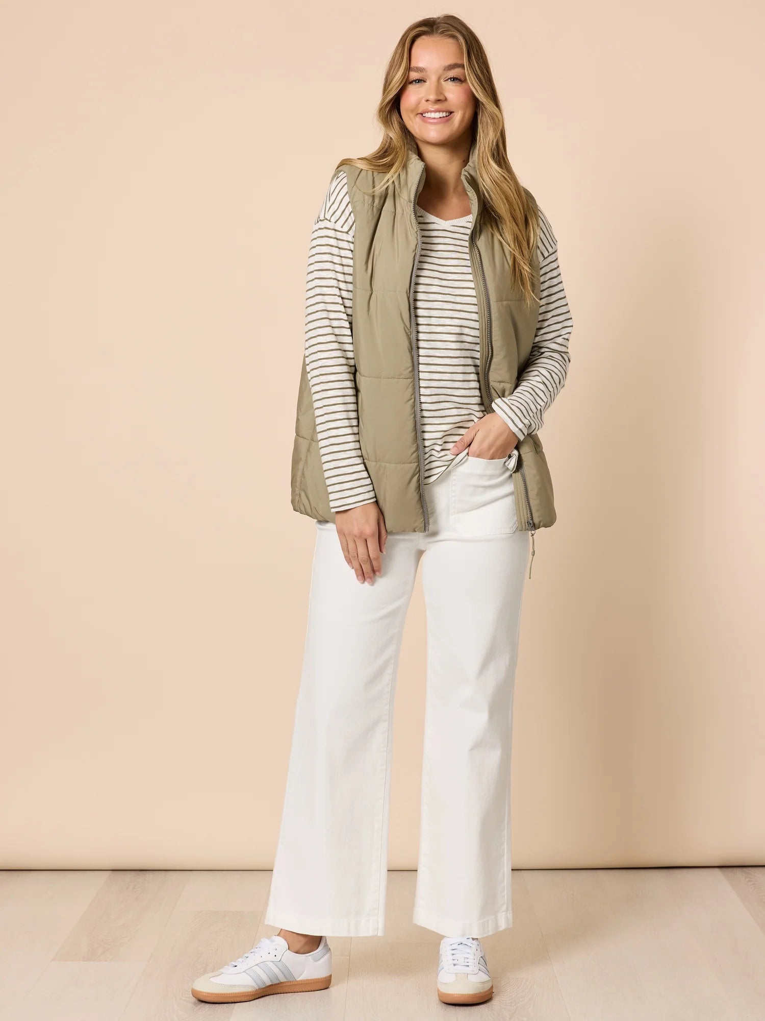 Image of Woman wearing a Threadz striped shirt, green vest, and white pants against a beige background