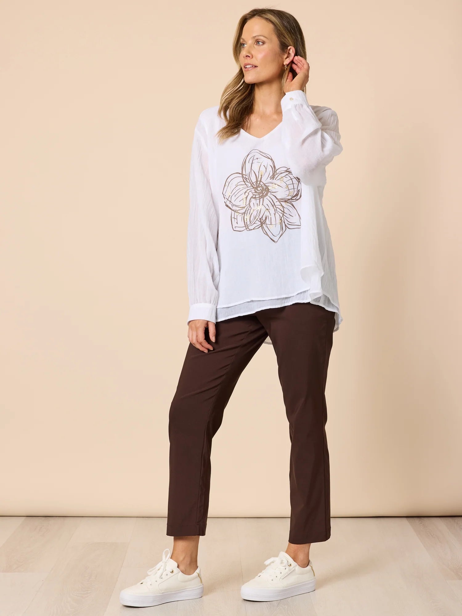 Woman wearing a white shirt with a floral design and Threadz brown pants on a beige background