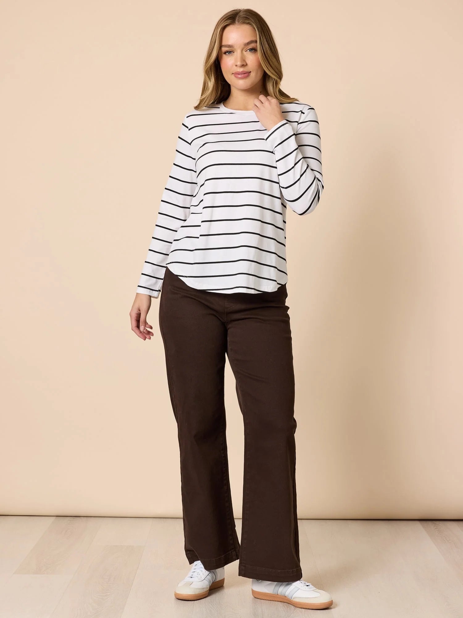 Image of A woman modeling a white long-sleeve top with thin black horizontal stripes and Threadz dark brown wide-leg pants, standing against a solid beige studio backdrop.