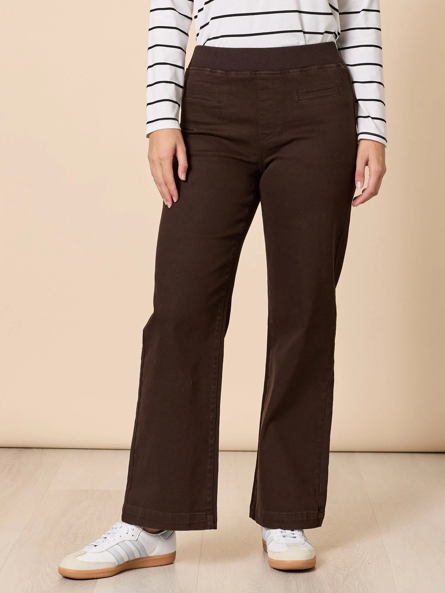 Image of A woman modeling a white long-sleeve top with thin black horizontal stripes and Threadz dark brown wide-leg pants, standing against a solid beige studio backdrop.