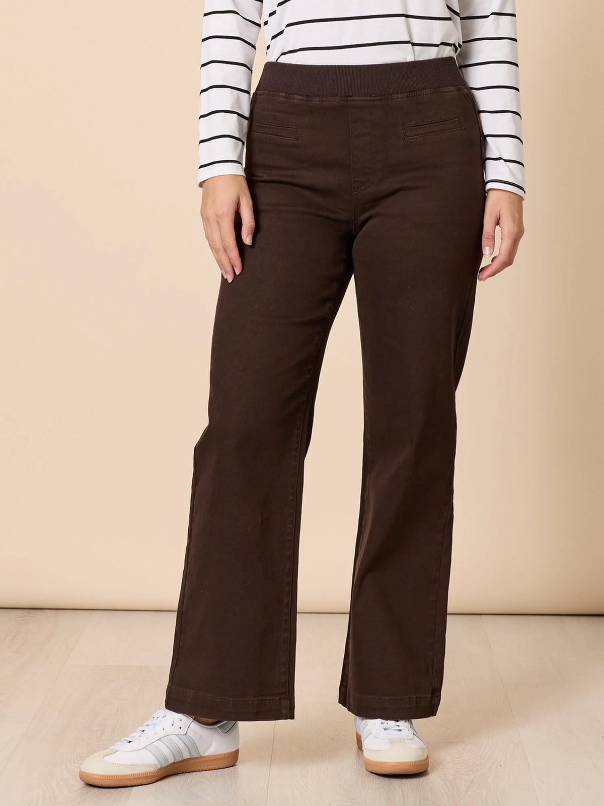 A woman modeling a white long-sleeve top with thin black horizontal stripes and Threadz dark brown wide-leg pants, standing against a solid beige studio backdrop.