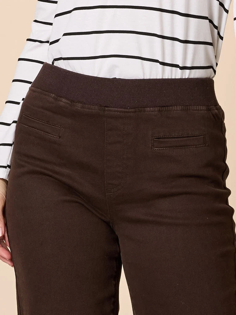 A woman modeling a white long-sleeve top with thin black horizontal stripes and Threadz dark brown wide-leg pants, standing against a solid beige studio backdrop.