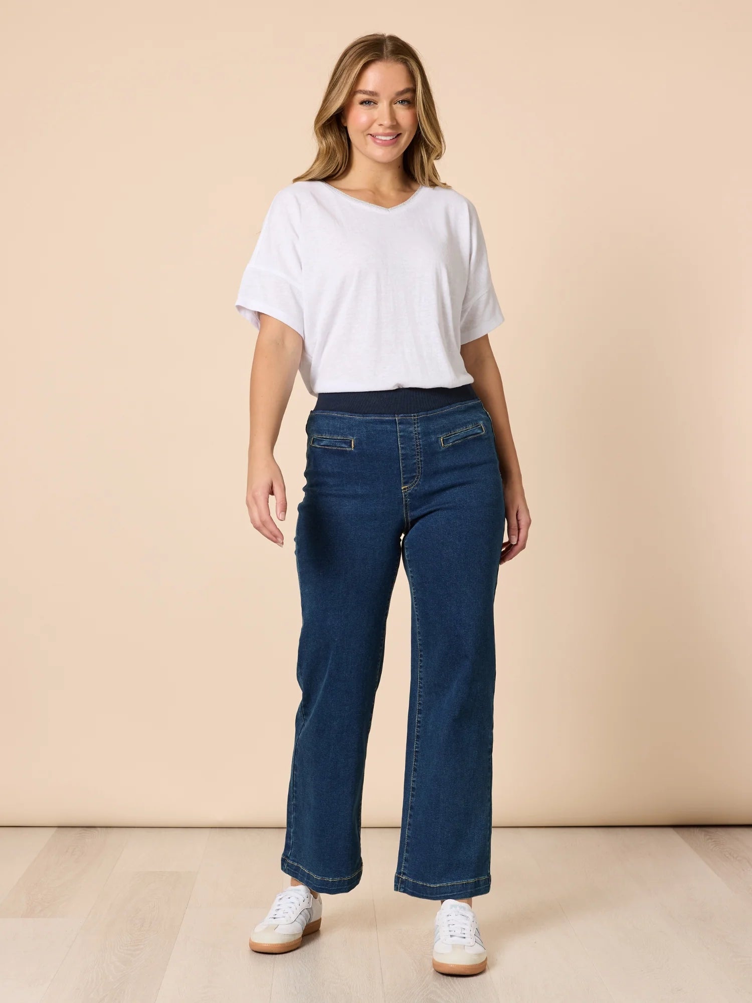 Image of A woman modeling a pair of Threadz dark blue wide-leg jeans with a white t-shirt and white sneakers, standing against a solid beige studio background.