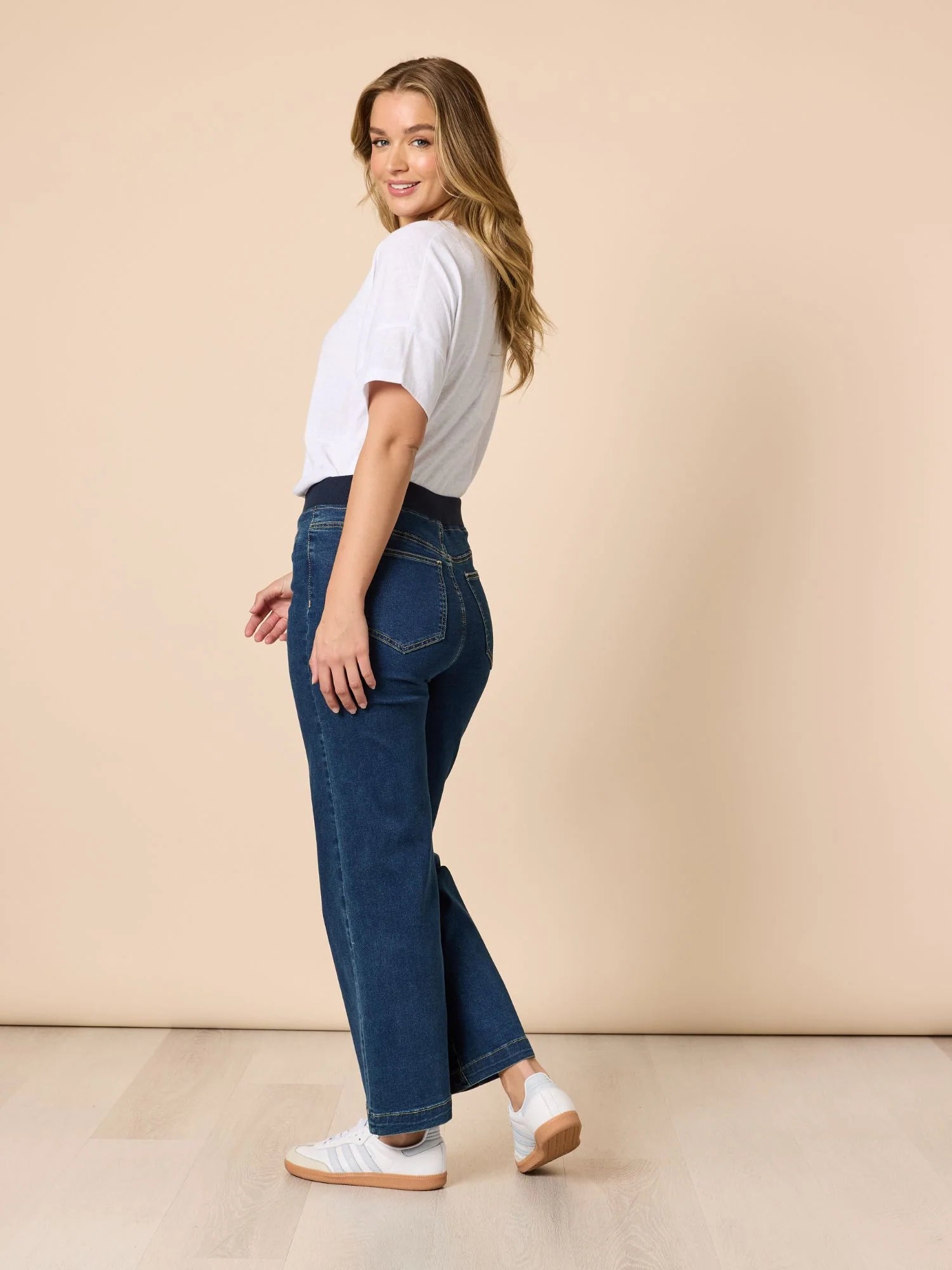 Image of A woman wearing Threadz dark blue high-waisted straight-leg jeans, a white t-shirt, and white sneakers, standing in a studio with a beige background.