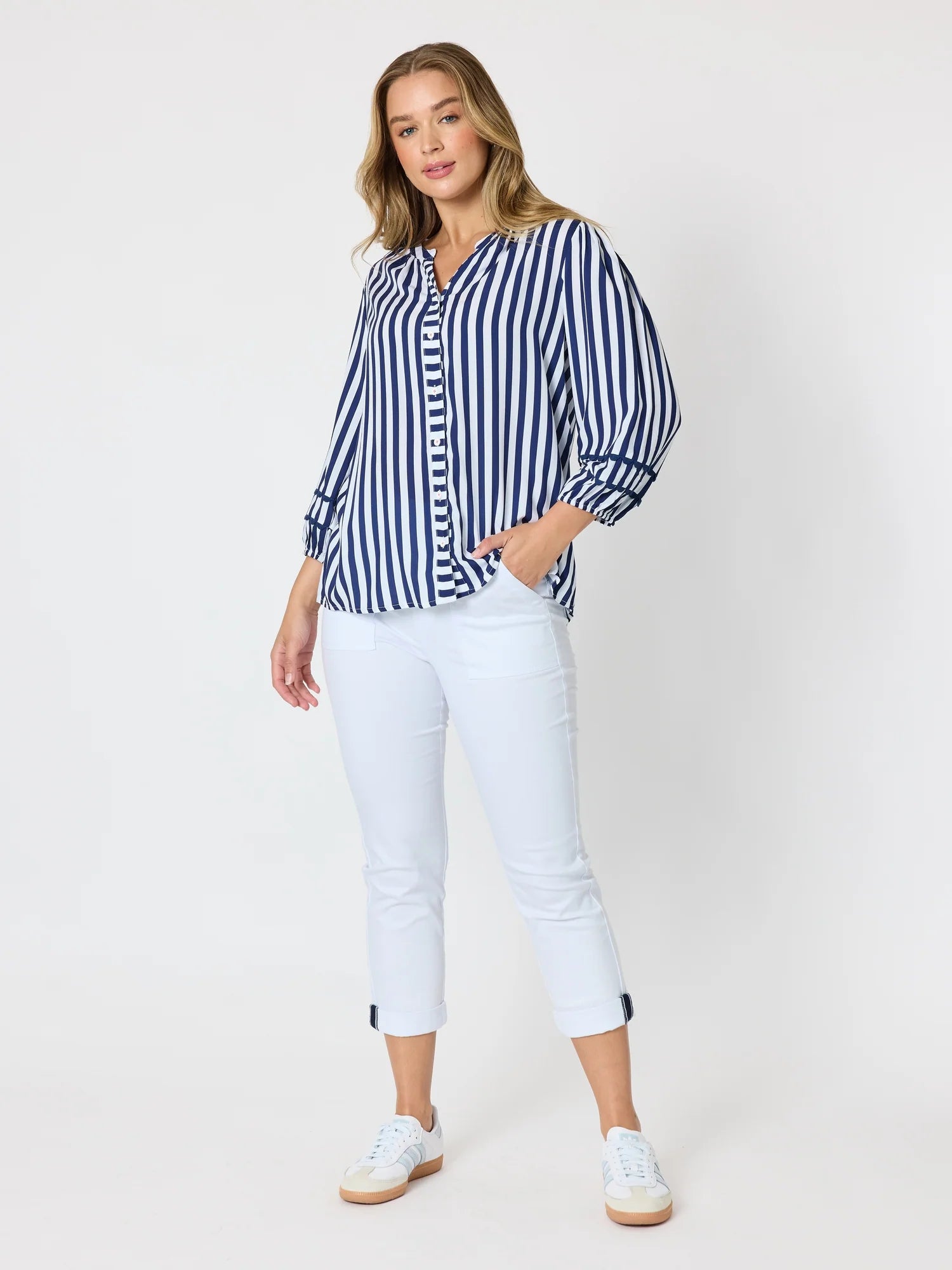 Image of A woman modeling a Threadz blue and white vertically striped button-up shirt, paired with white cropped pants and white sneakers, against a plain white studio background.