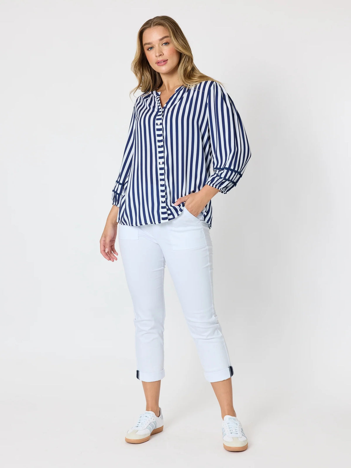 A woman modeling a Threadz blue and white vertically striped button-up shirt, paired with white cropped pants and white sneakers, against a plain white studio background.