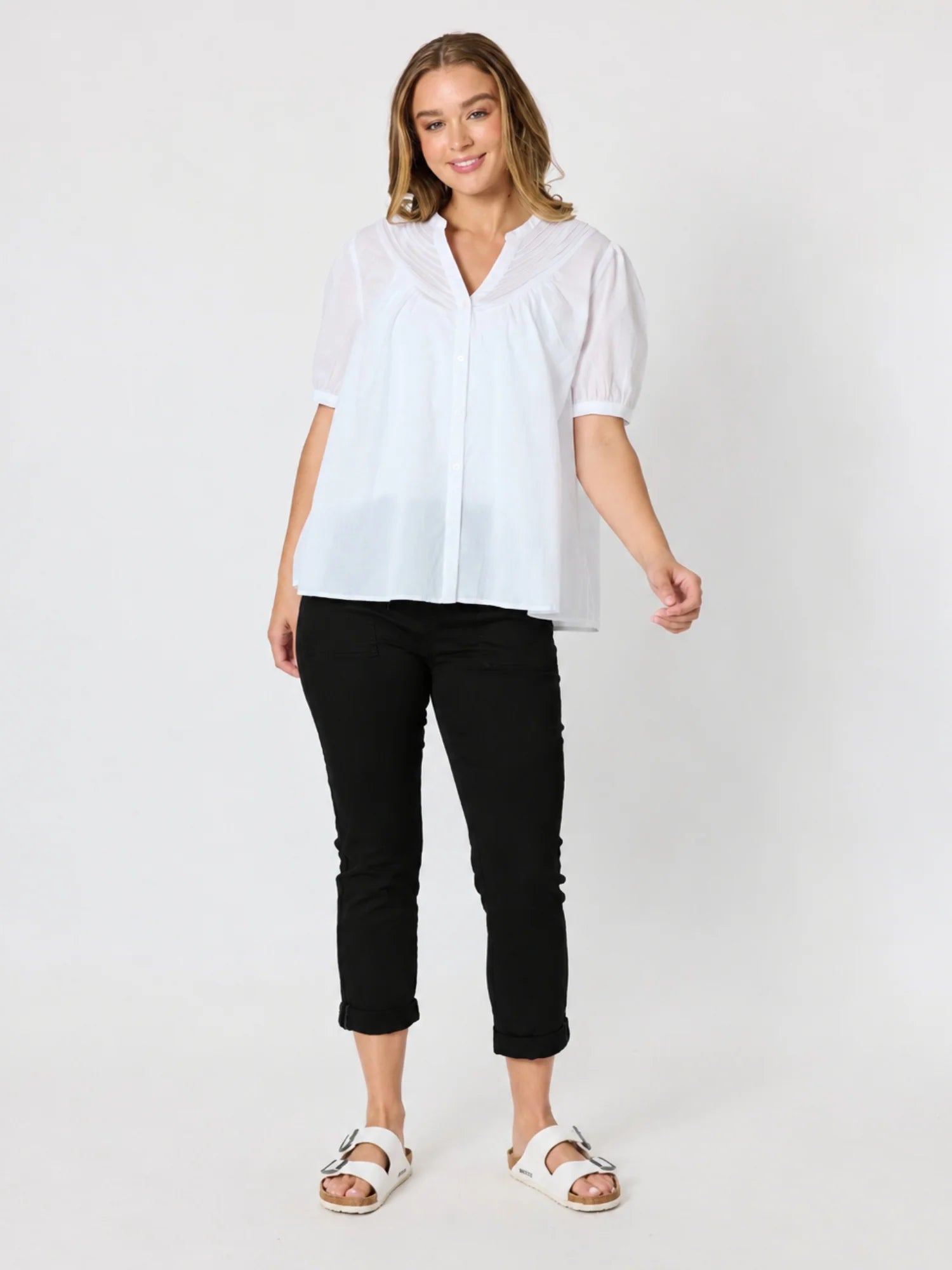 Image of A woman modeling a white short-sleeve blouse with a V-neck and sheer puff sleeves, paired with Threadz black cropped pants and white sandals, against a light gray studio background.