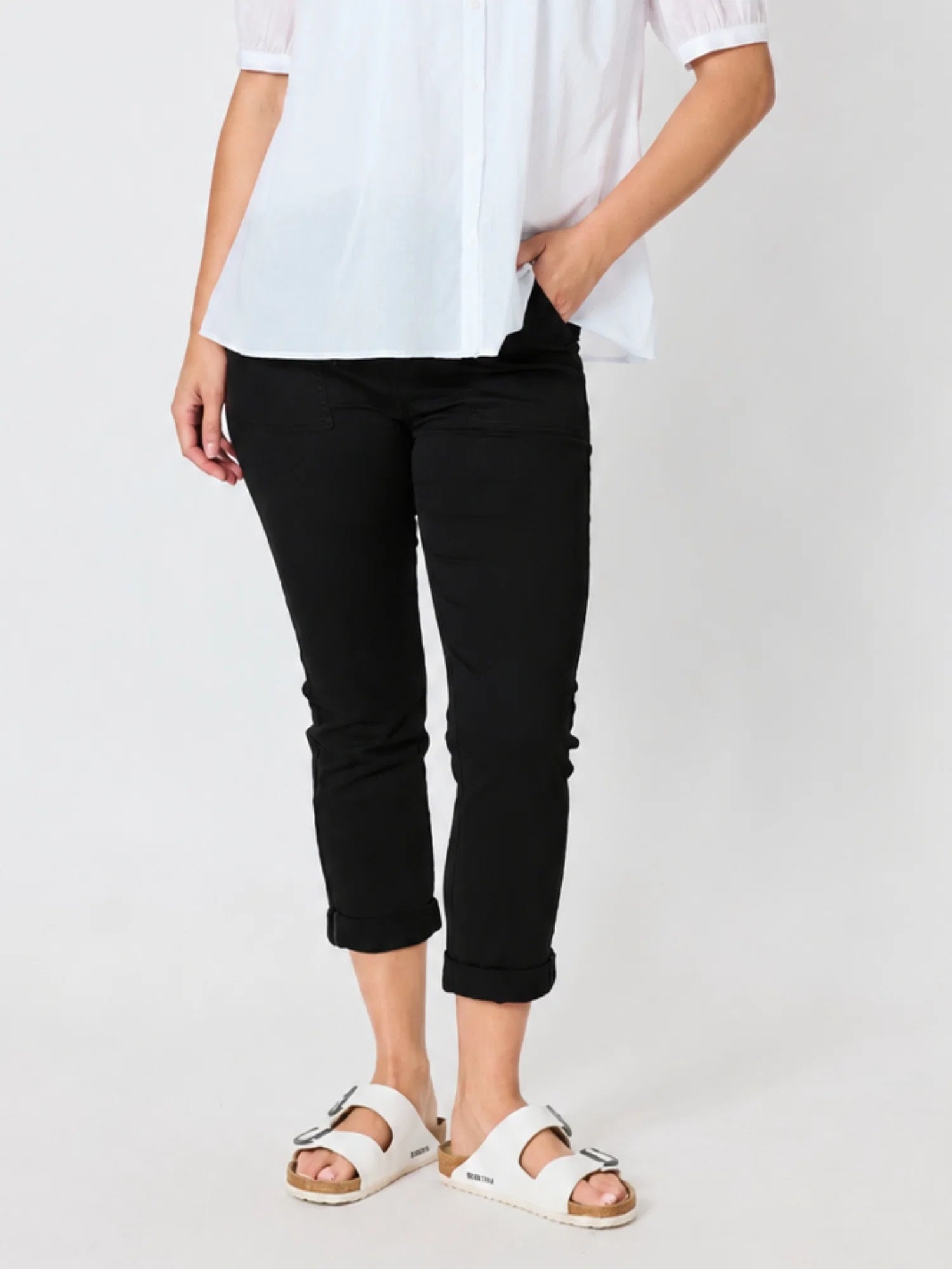 Image of A model wearing Threadz black cropped pants, a white blouse, and white slide sandals, standing against a plain light gray studio background.