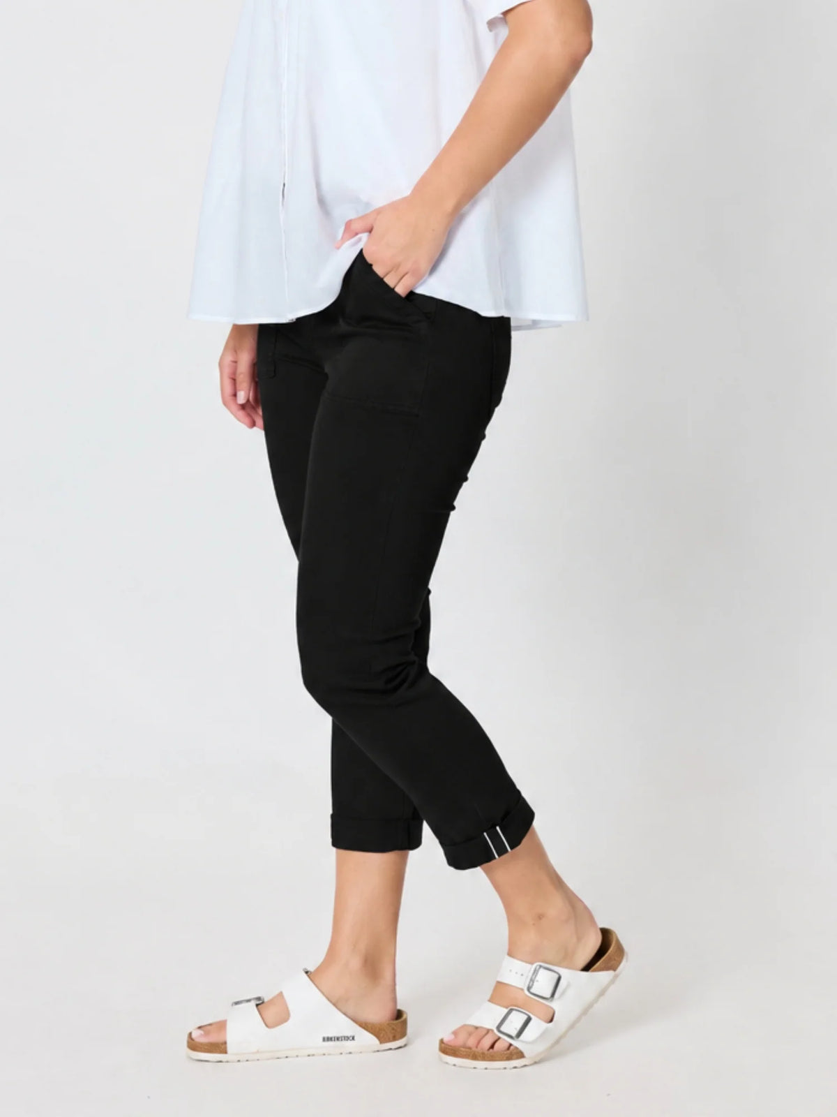 Woman wearing Threadz black cropped pants with rolled cuffs, a loose white top, and white slide sandals, photographed against a plain light gray studio background.