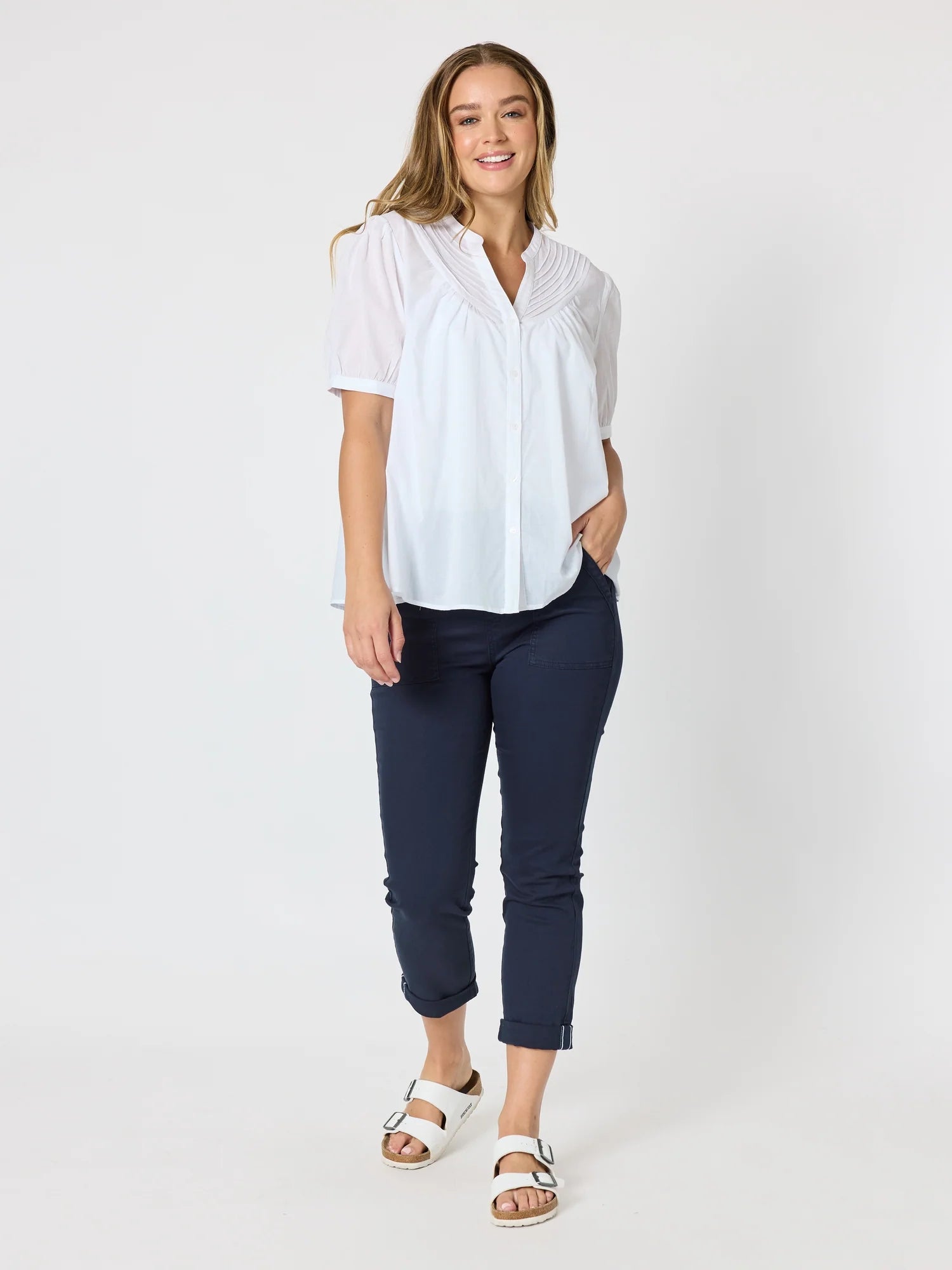 Image of A woman modeling a white short-sleeve button-up blouse with pleated detailing on the chest, paired with Threadz navy cropped pants and white slide sandals, against a plain white studio background.
