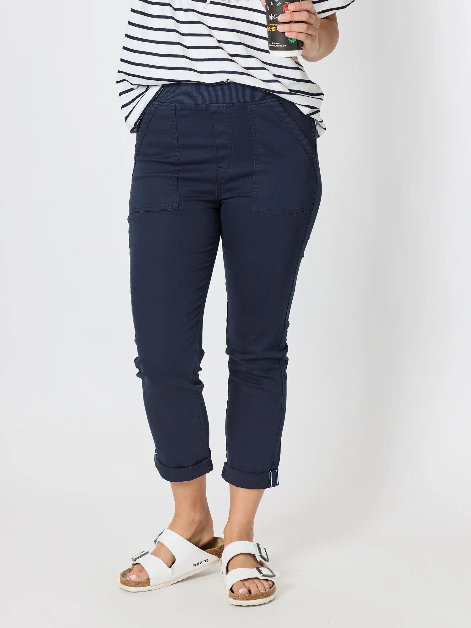 Image of Woman wearing Threadz navy blue cropped pants with a striped shirt and white slide sandals, holding a drink against a plain light background.