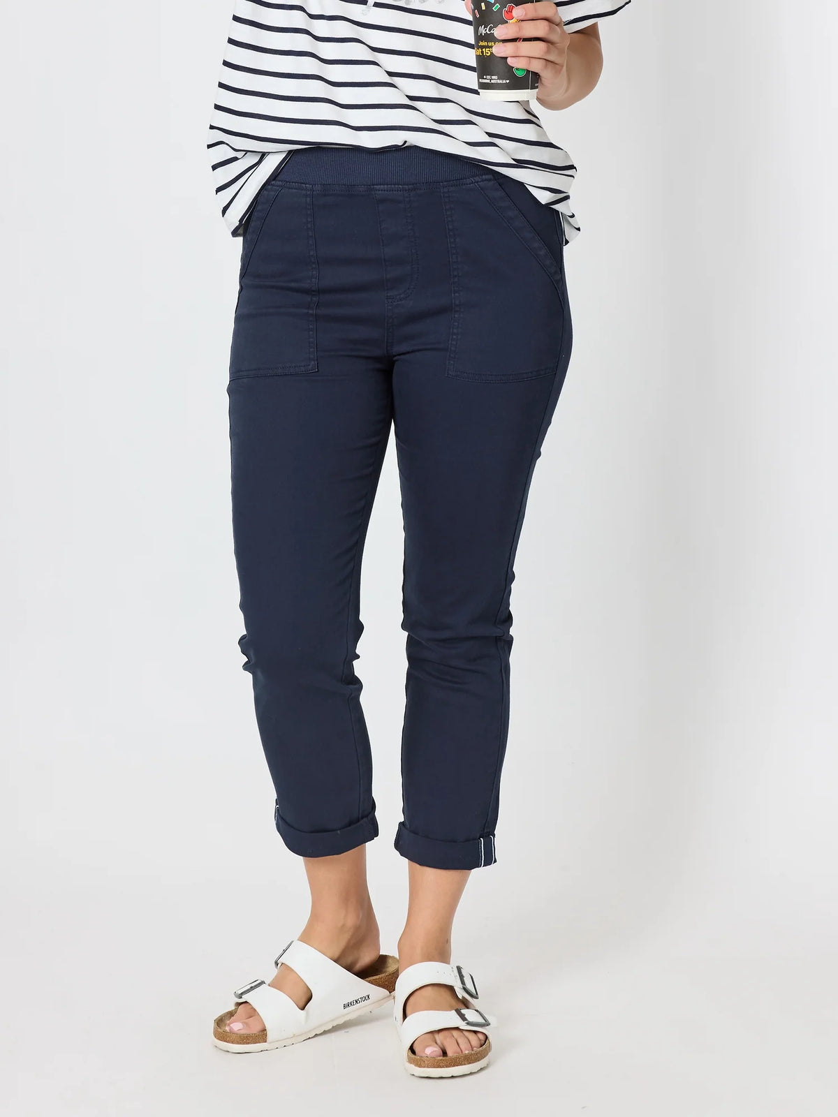 Woman wearing Threadz navy blue cropped pants with a striped shirt and white slide sandals, holding a drink against a plain light background.