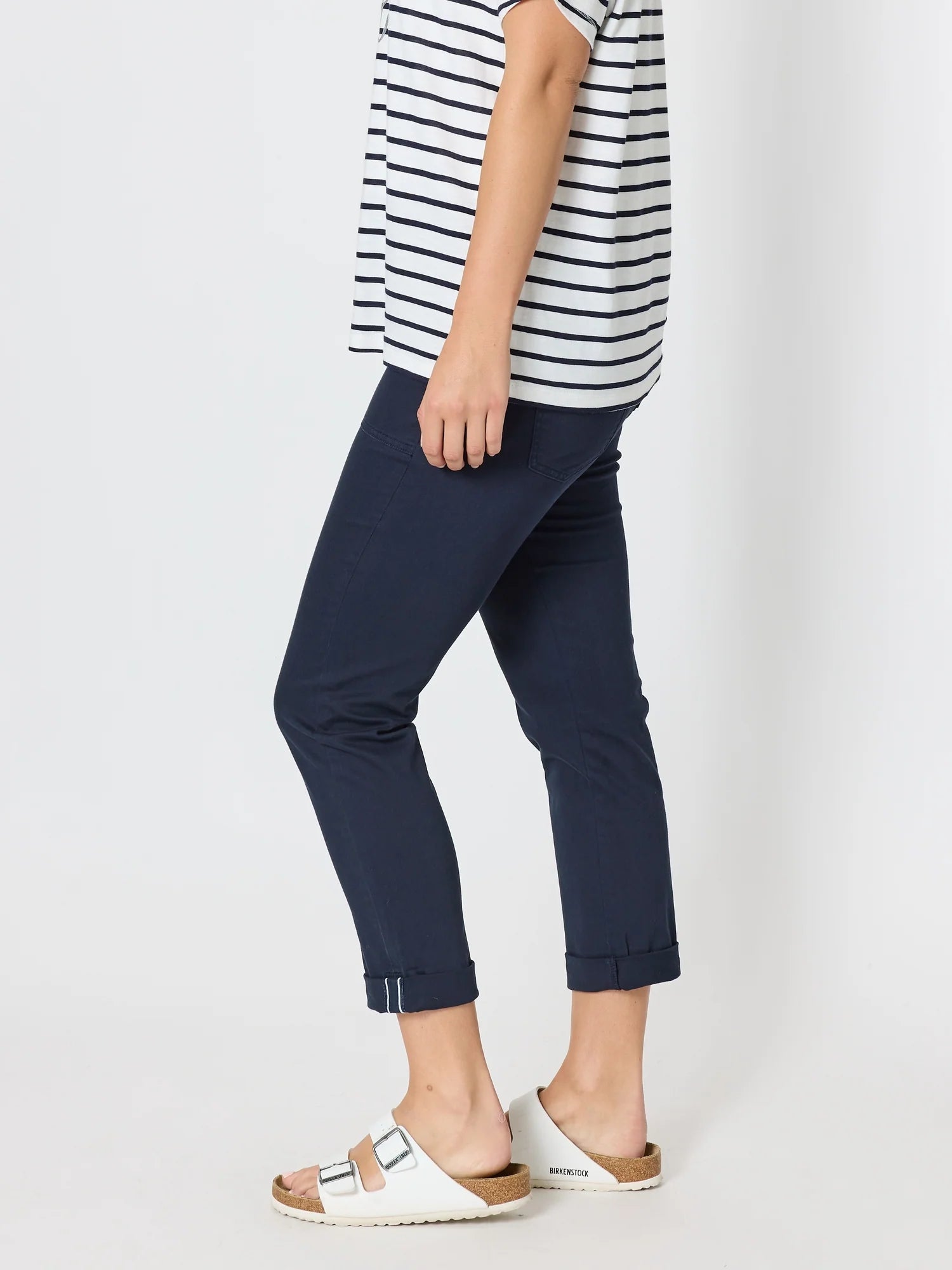 Image of A woman shown from the waist down wearing Threadz navy blue cropped pants with rolled cuffs, a navy and white striped shirt, and white slide sandals on a light gray studio background.