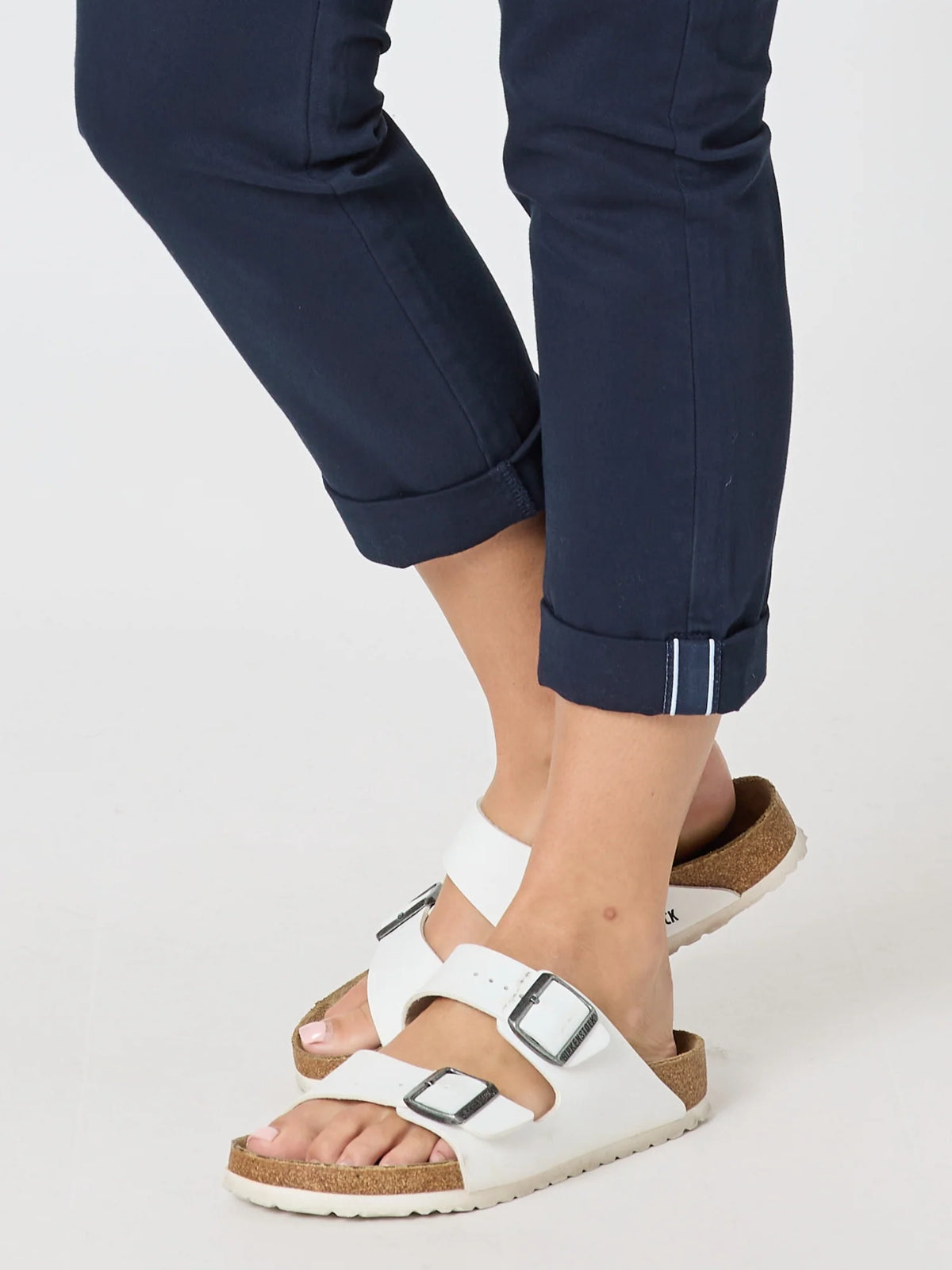 White double-strap slide sandals with adjustable buckles and a cork-look midsole, shown worn with Threadz  navy cuffed pants on a light gray studio background.