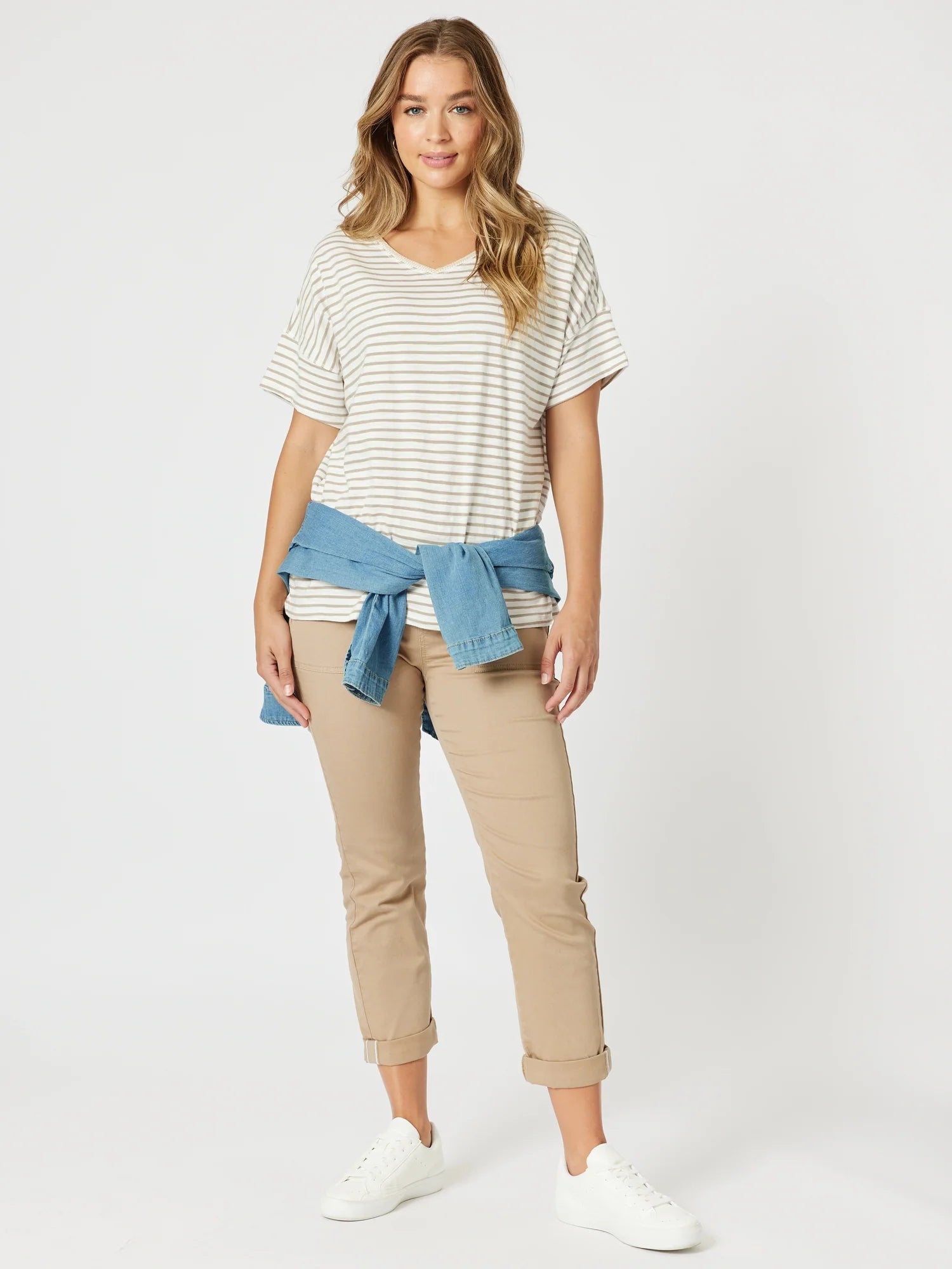Image of A woman modeling a casual outfit consisting of a Threadz cream and gray striped short-sleeve t-shirt, a blue denim shirt tied around her waist, beige cropped pants, and white sneakers, against a light gray studio background.