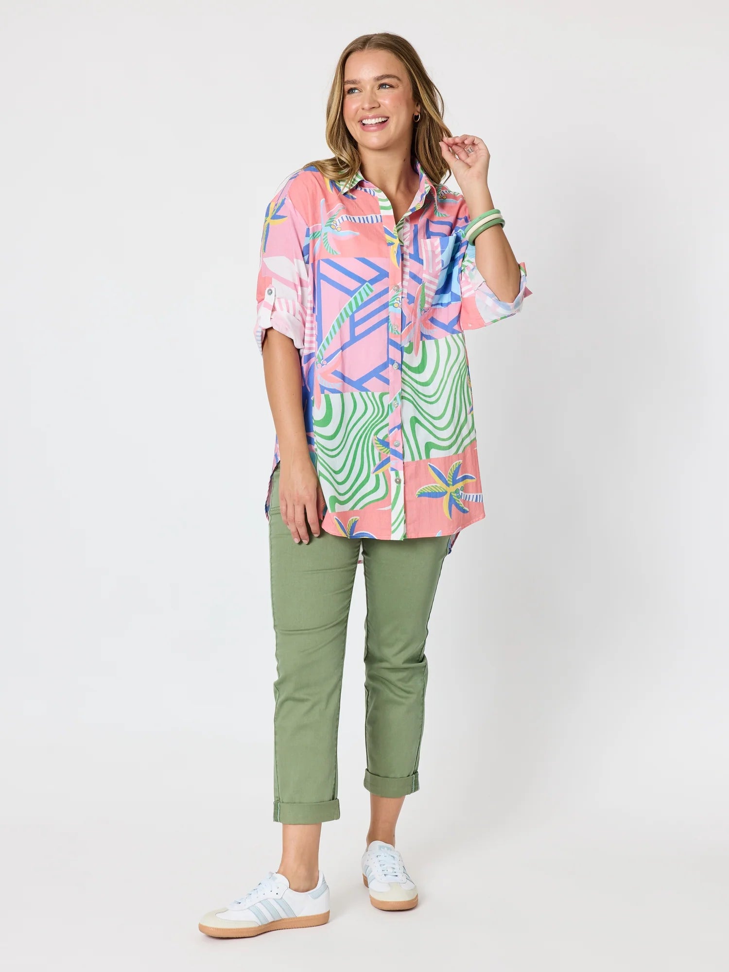 Image of A woman modeling a short-sleeve button-up shirt with a vibrant pink, green, and blue tropical and geometric print, paired with Threadz olive green cropped pants and white sneakers, against a plain white studio background.