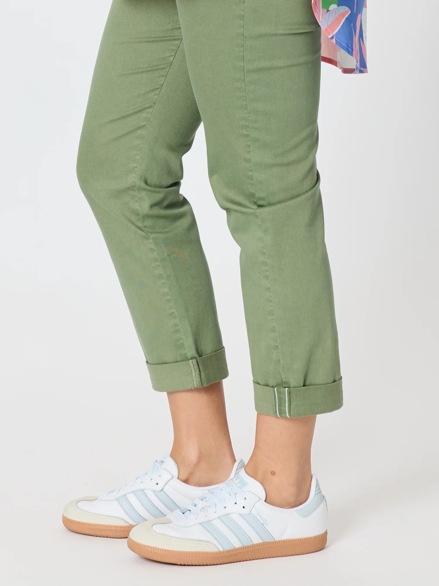 Image of A woman wearing Threadz sage green cropped pants with rolled cuffs and white low-top sneakers with light blue stripes, shown from the waist down on a light gray studio background.