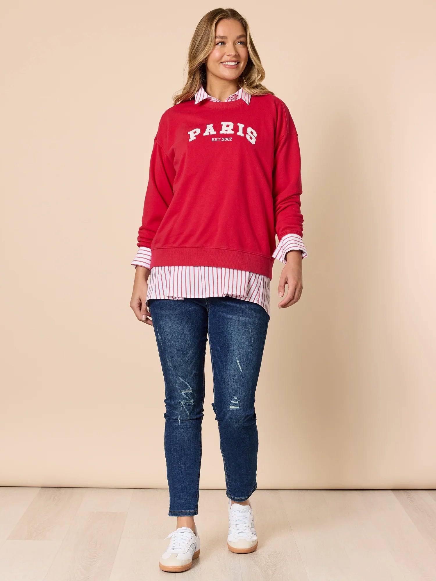 Woman wearing a Threadz red sweatshirt with 'Paris' text, blue jeans, and white sneakers on a beige background