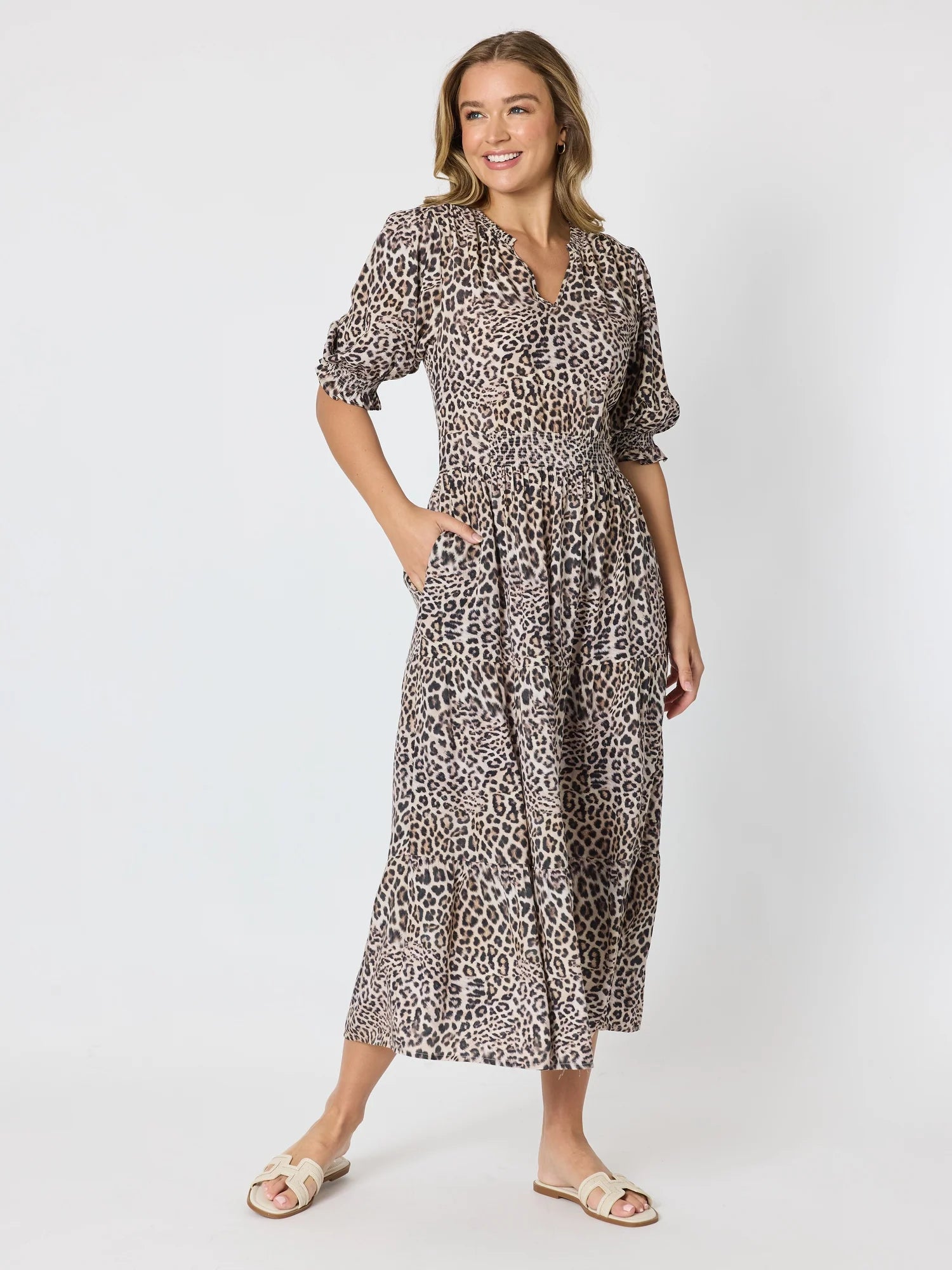 A woman modeling a Threadz midi-length leopard print dress with puff sleeves, a V-neckline, and side pockets, paired with beige slide sandals against a light gray studio background.