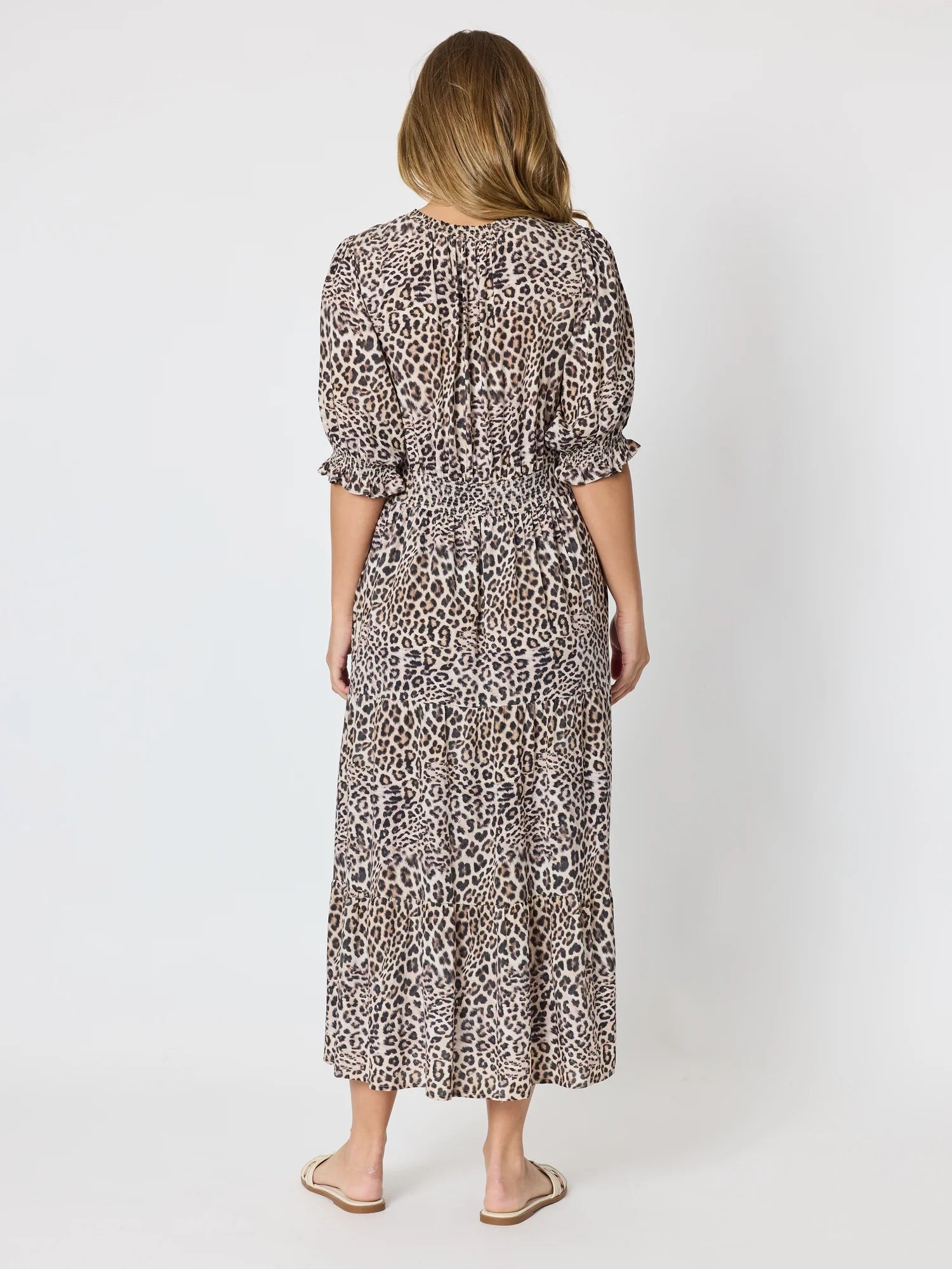 Image of A woman viewed from the back wearing a Threadz midi-length leopard print dress with puff sleeves and a smocked waist, paired with beige flat sandals, against a plain light gray studio background.
