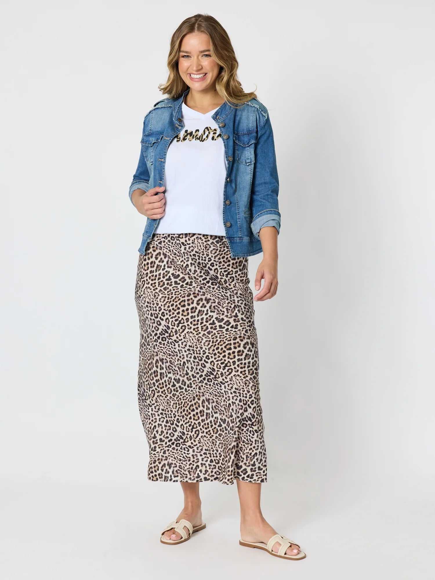 A woman modeling a Threadz leopard-print midi skirt, styled with a white t-shirt, a blue denim jacket, and beige slide sandals, against a light gray studio background.