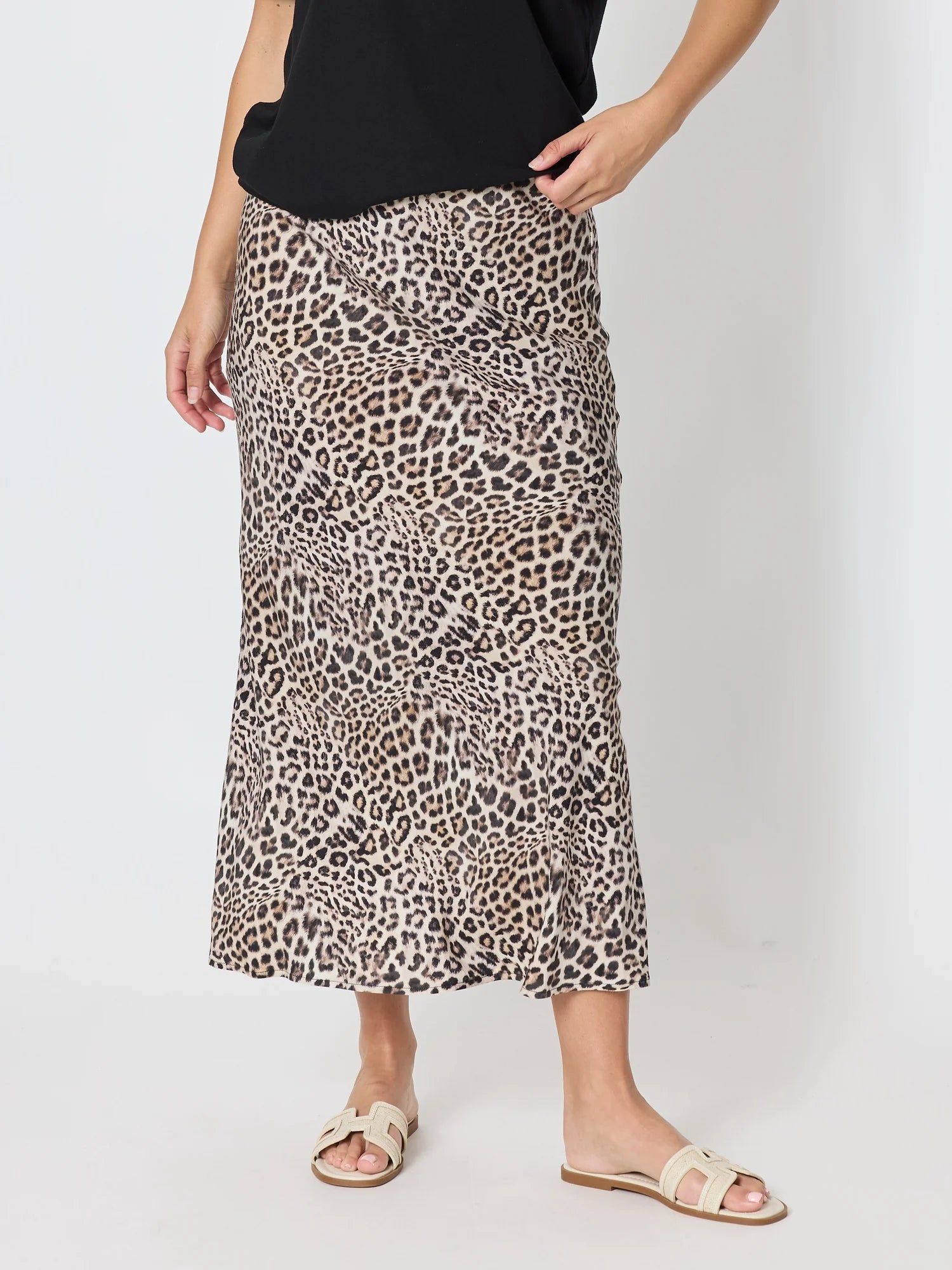 A woman wearing a Threadz leopard print midi skirt with a black top and beige slide sandals, standing against a plain light gray studio background.