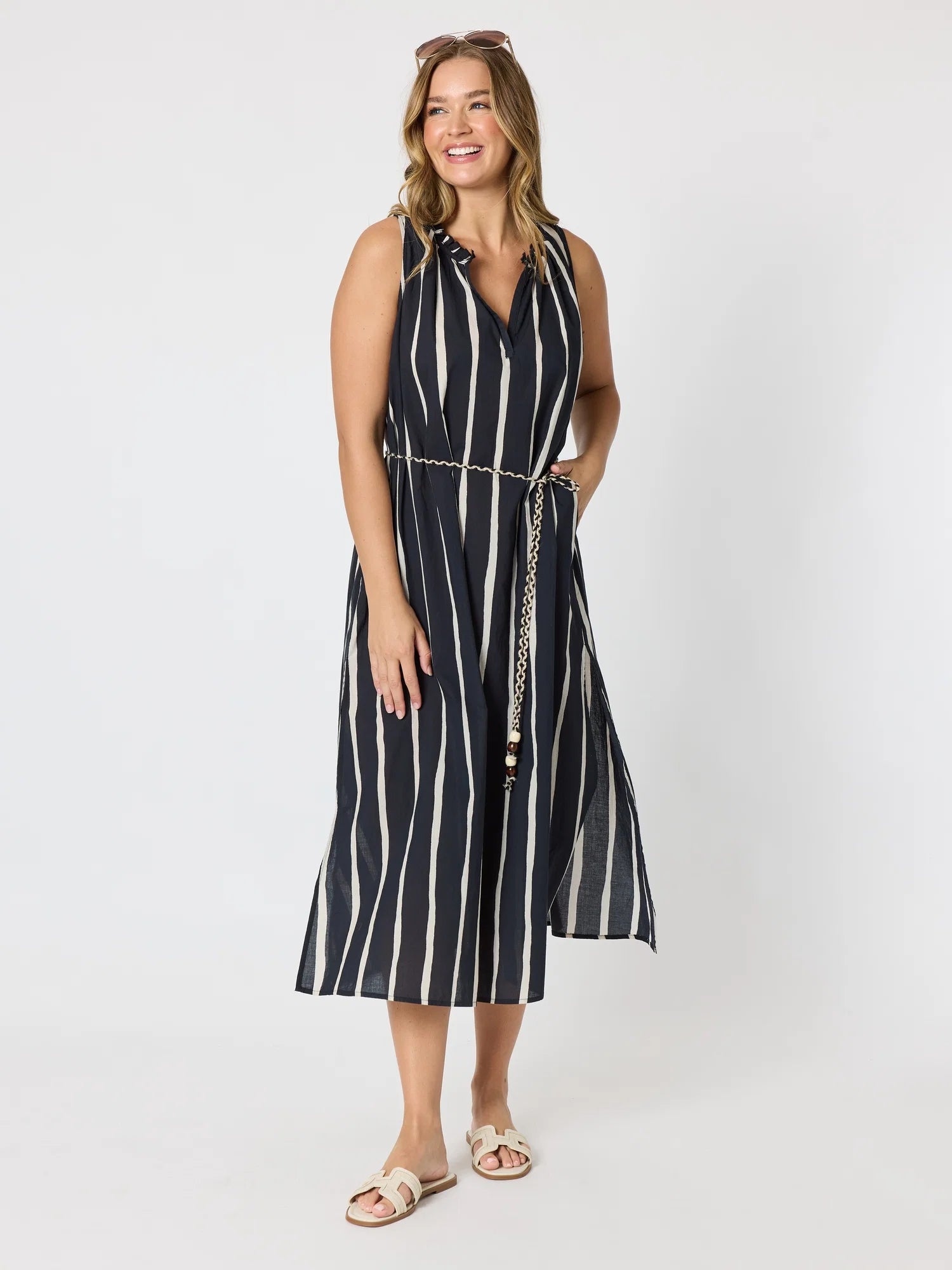 Image of A woman modeling a Threadz sleeveless midi dress with vertical black and white stripes, featuring a V-neckline, a matching striped tie belt with a chain detail, and flat beige sandals, set against a plain light gray studio background.