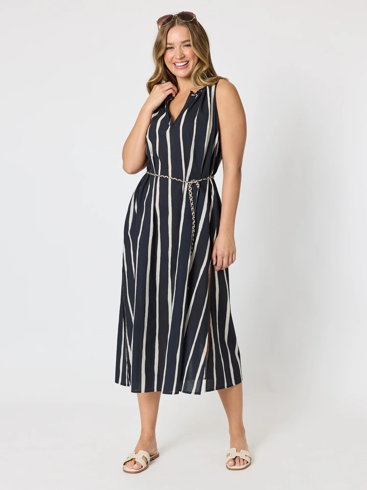 Image of A woman modeling a Threadz sleeveless midi dress with vertical navy and off-white stripes, featuring a V-neckline, a matching thin chain belt at the waist, and light beige slide sandals, set against a plain white studio background.