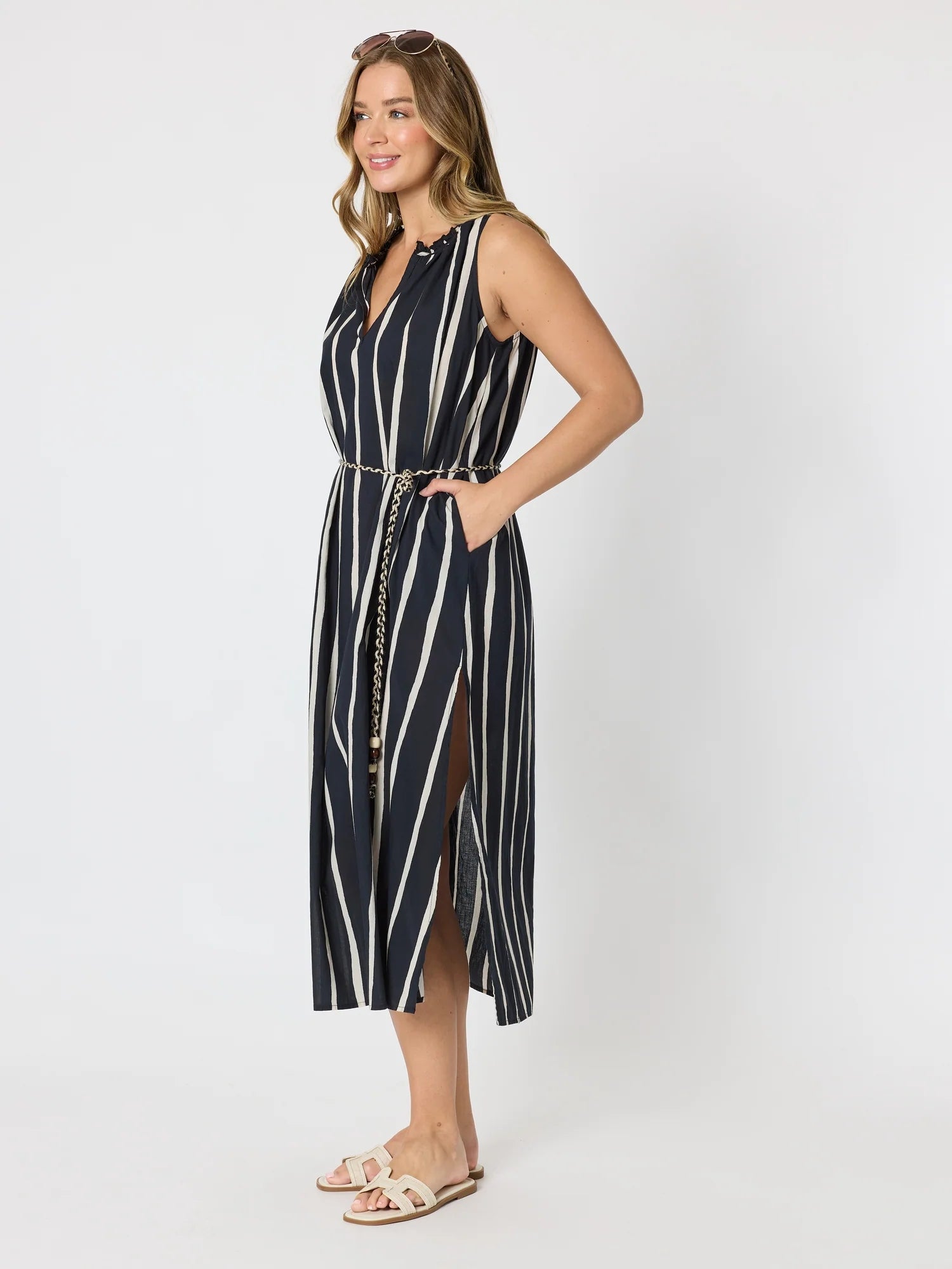 Image of A woman modeling a Threadz sleeveless midi dress with vertical navy and white stripes, a V-neckline, side pockets, and a matching tie belt with a gold chain detail, set against a plain light gray studio background.