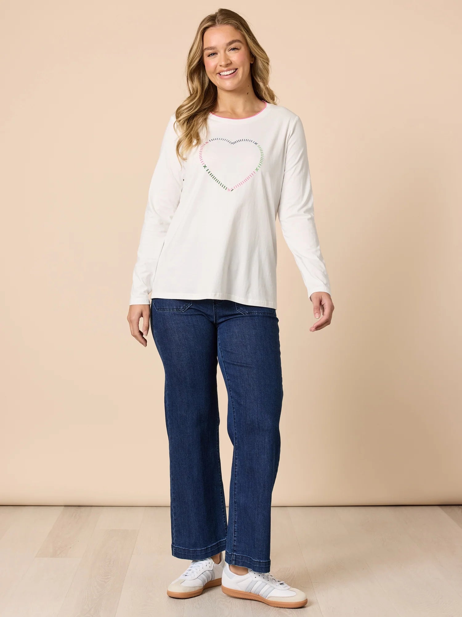 Image of Woman wearing a Threadz white long-sleeve top with a heart design and blue jeans on a beige background