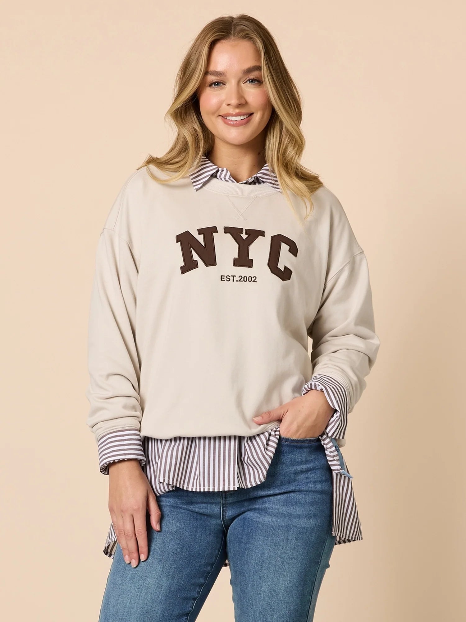 Woman wearing a Threadz beige sweatshirt with 'NYC' text, striped shirt, and jeans on a beige background