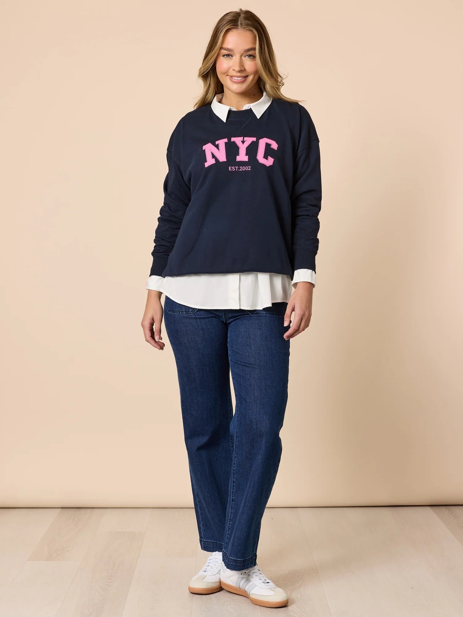 Image of Woman wearing a Threadz navy sweatshirt with 'NYC' text, white shirt, and blue jeans on a beige background
