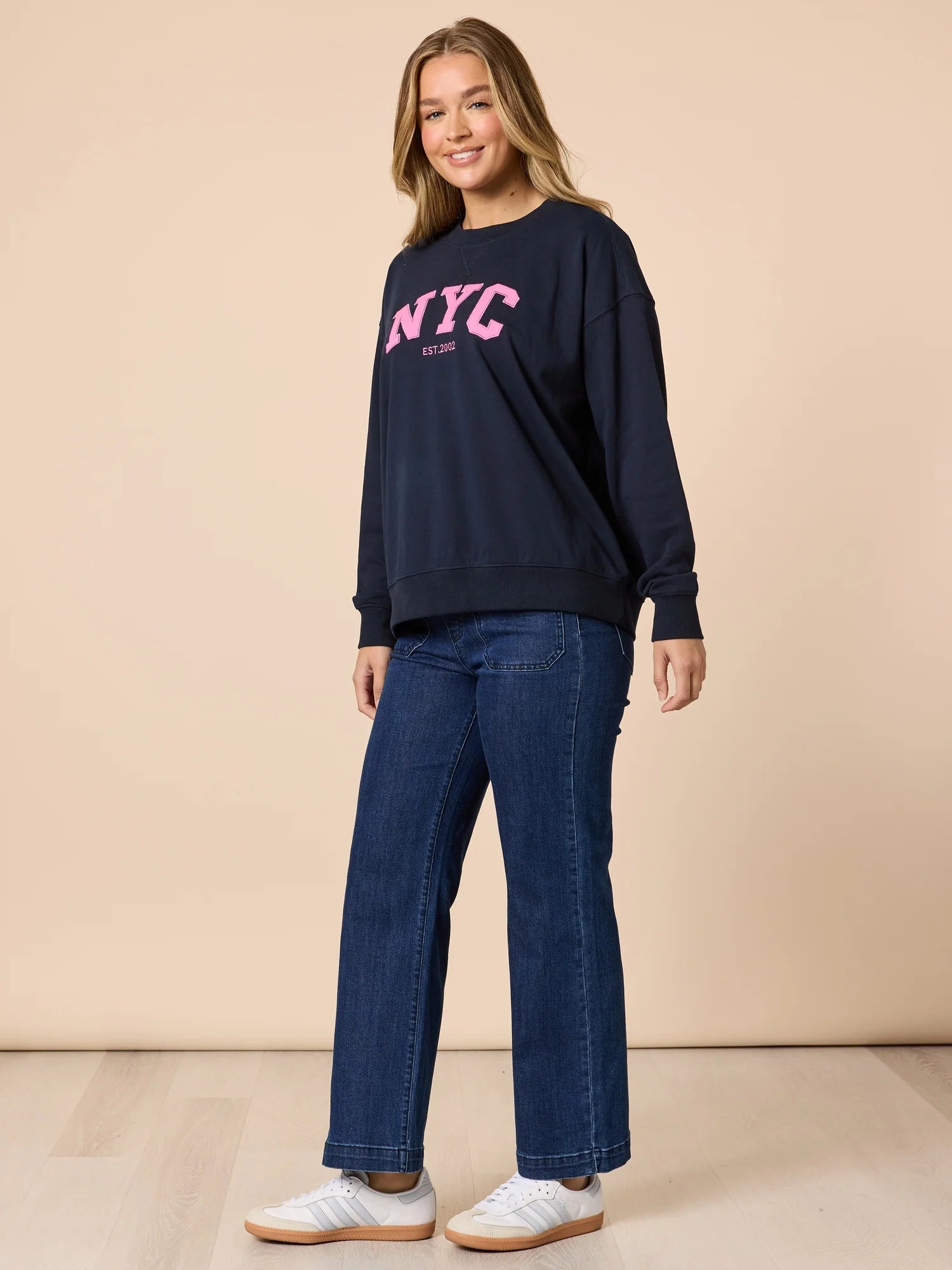 Woman wearing a Threadz navy sweatshirt with 'NYC' in pink and blue jeans on a beige background