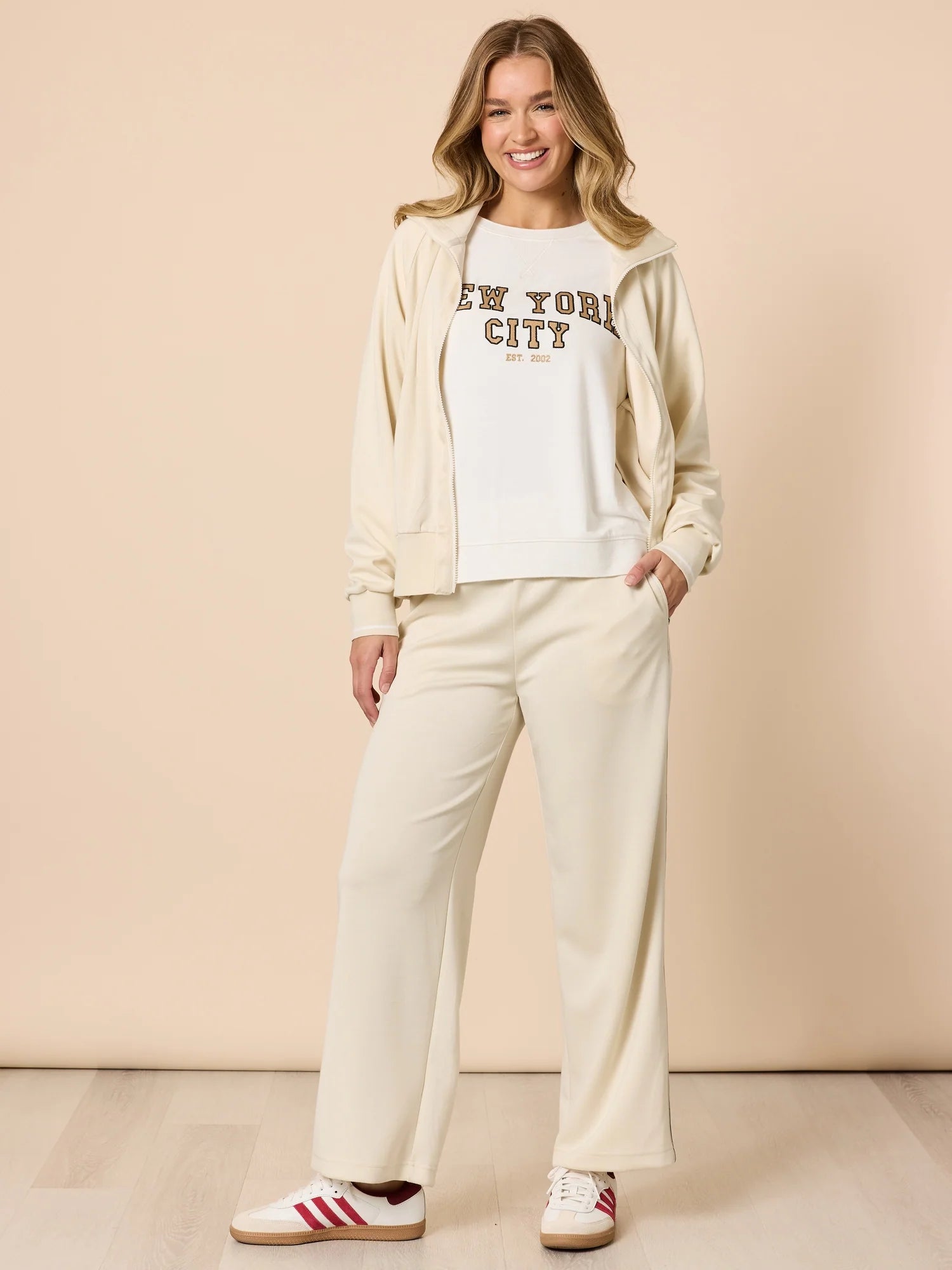 Woman wearing a Threadz 'New York City' sweatshirt with cream pants and jacket on a beige background