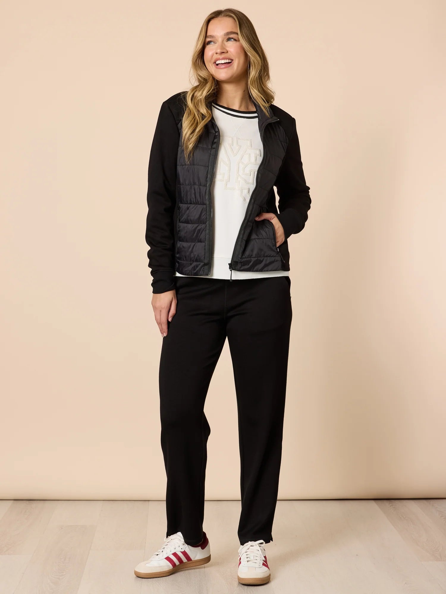 Woman wearing a black puffer jacket, white shirt, Threadz black pants, and red Adidas sneakers on a beige background