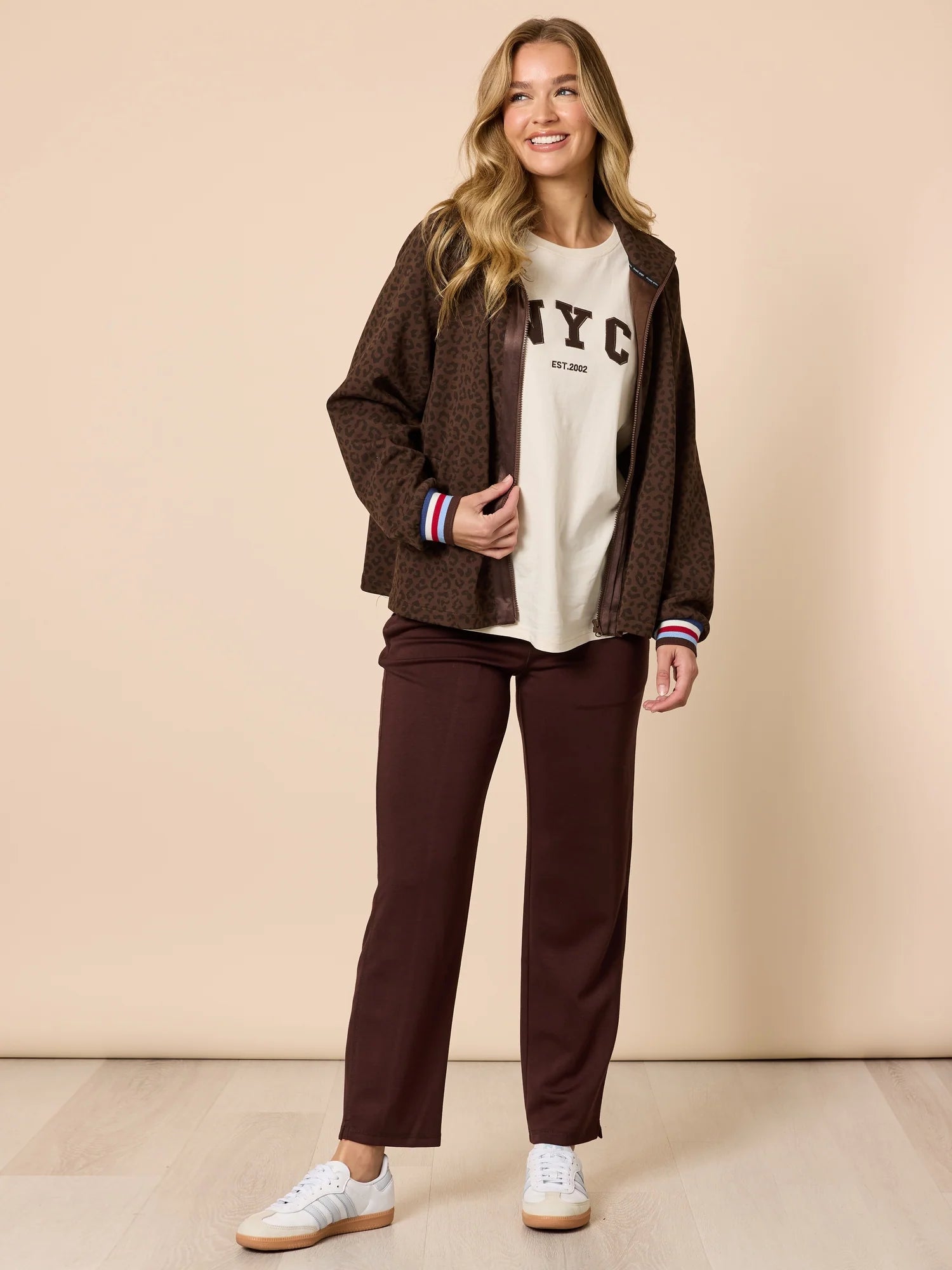 Image of Woman wearing a brown jacket, white t-shirt with 'NYC' print, and Threadz brown pants on a beige background