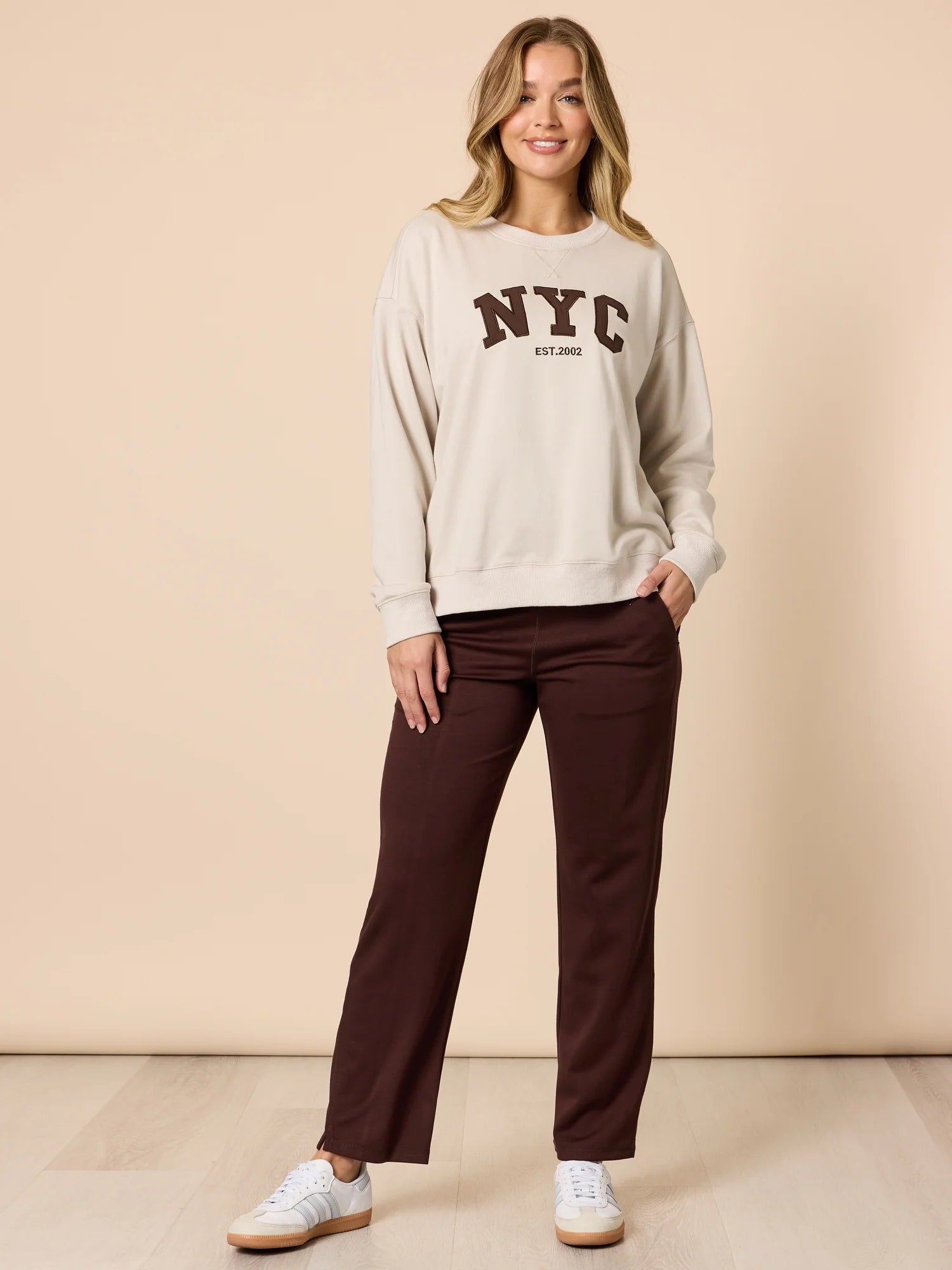 Image of Woman wearing a beige sweatshirt with 'NYC' text and Threadz brown pants on a beige background