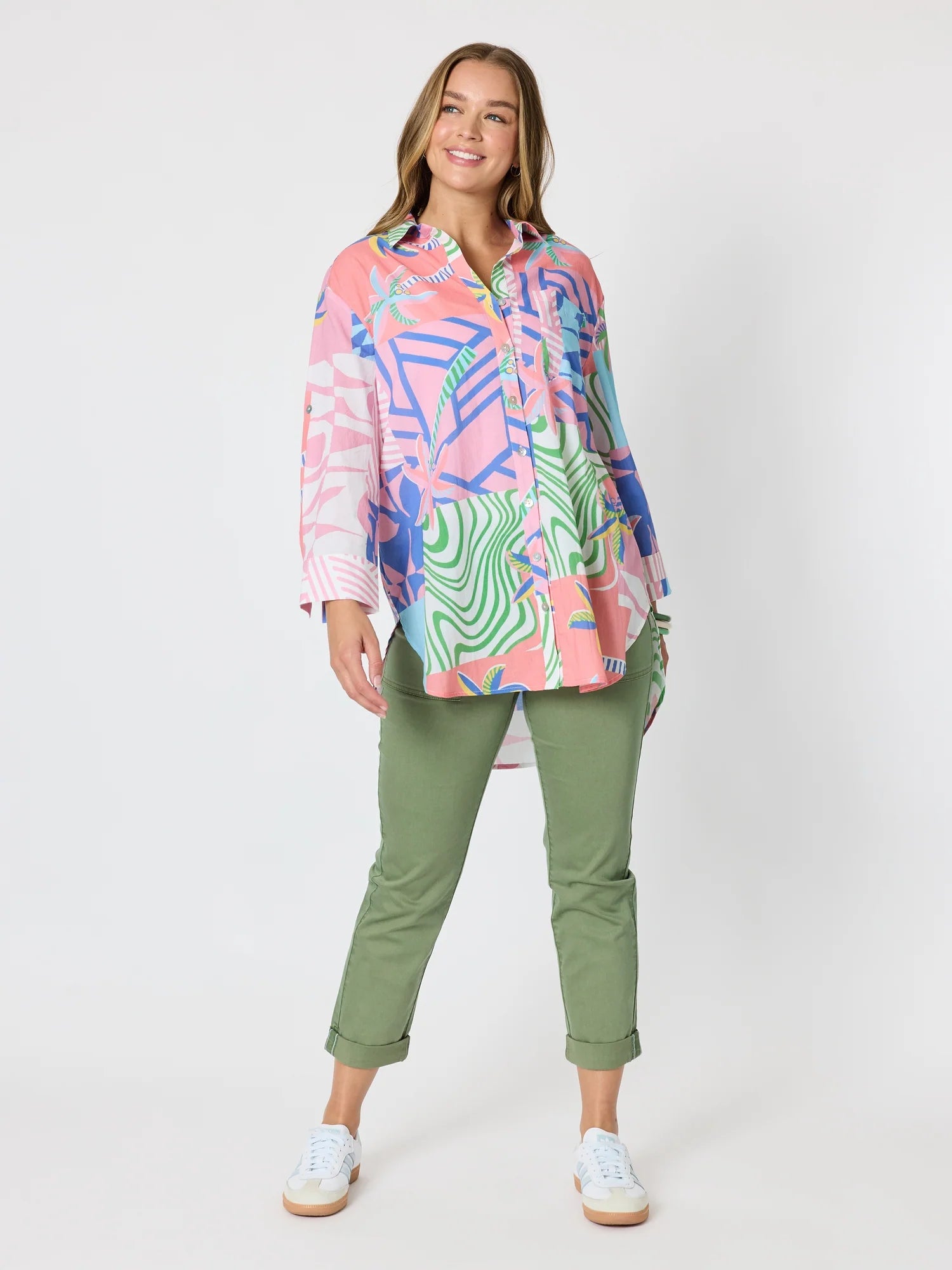 Image of A woman modeling a Threadz long-sleeve button-up shirt with a vibrant multicolor abstract print in pink, blue, green, and white, paired with olive green cropped pants and white sneakers, against a light gray studio background.