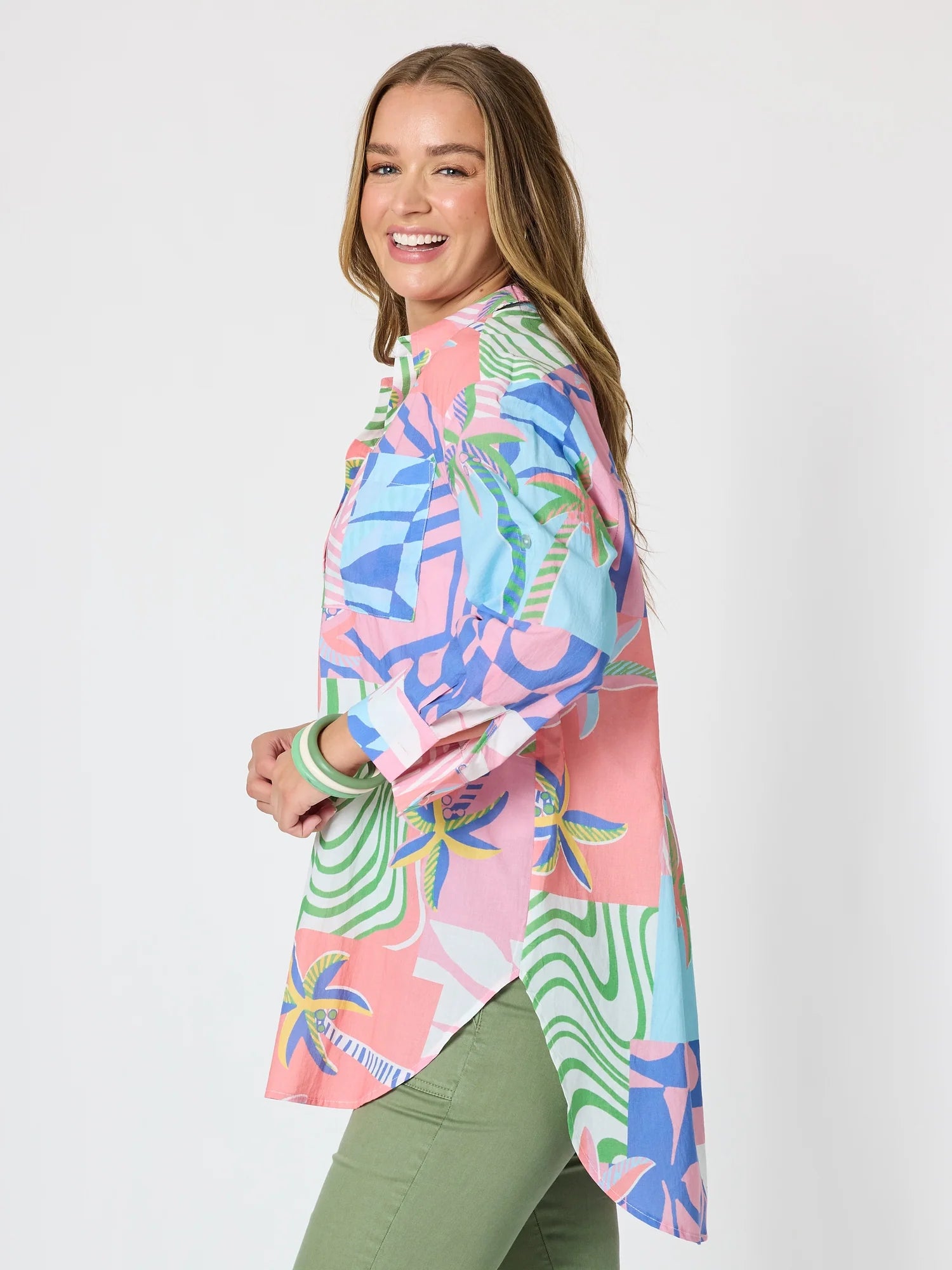 Image of A woman modeling a Threadz colorful tropical print button-up shirt with pink, blue, and green patterns, paired with olive green pants, against a light gray studio background.