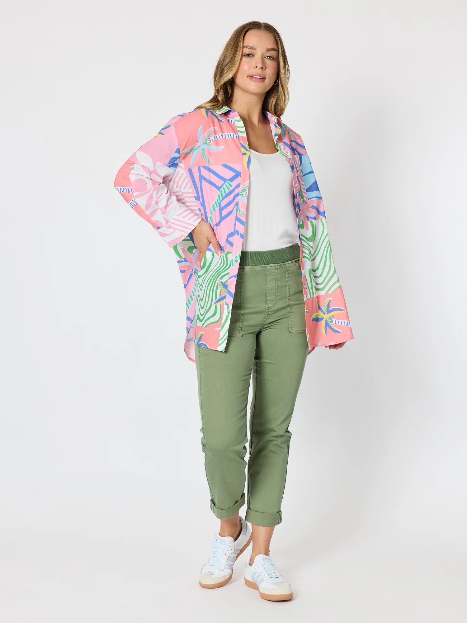A woman modeling a Threadz colourful patterned button-up shirt jacket over a white tank top and olive green cropped pants, paired with white sneakers, against a light gray studio background.
