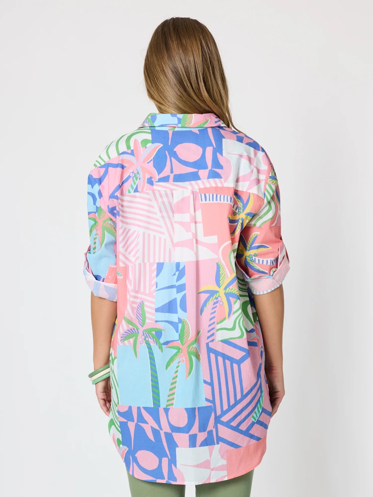 A woman viewed from the back wearing a Threadz colorful patchwork button-up shirt with a tropical and geometric print in pink, blue, green, and white, paired with olive green pants against a plain white background.