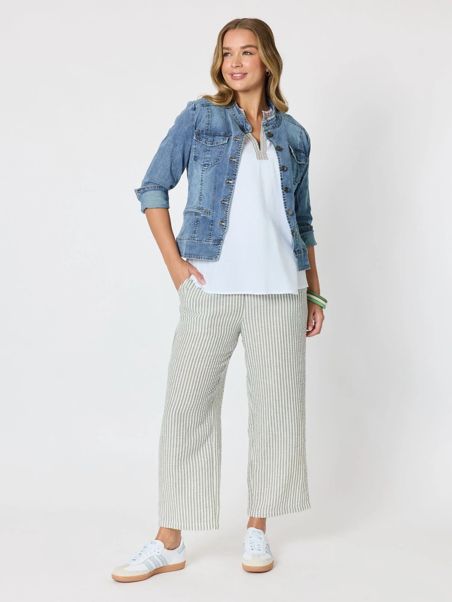Image of A woman modeling a casual outfit consisting of a light blue denim jacket, a white t-shirt, Threadz wide-leg striped pants, and white sneakers, photographed against a solid off-white studio background.