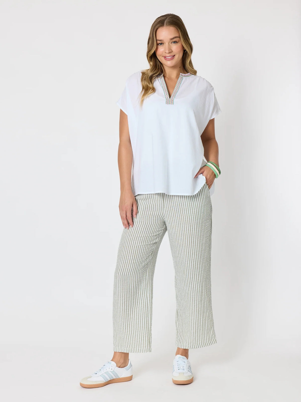 A woman modeling a white short-sleeve top with a split neckline and striped Threadz cropped pants in white and green, paired with white sneakers, against a plain white studio background.