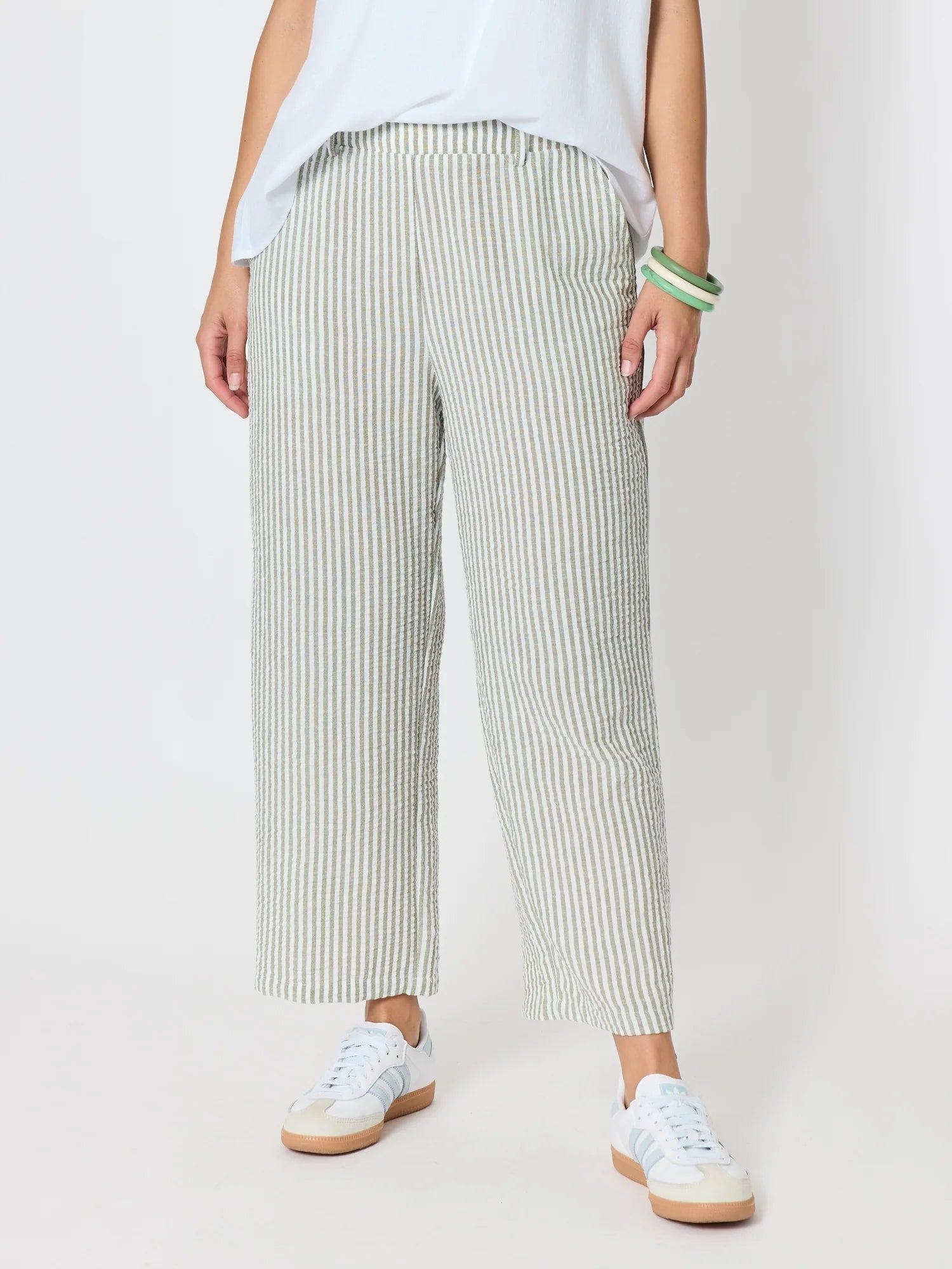 Image of A woman wearing Threadz light green and white vertically striped cropped trousers, styled with a white top and white sneakers against a plain light gray studio background.