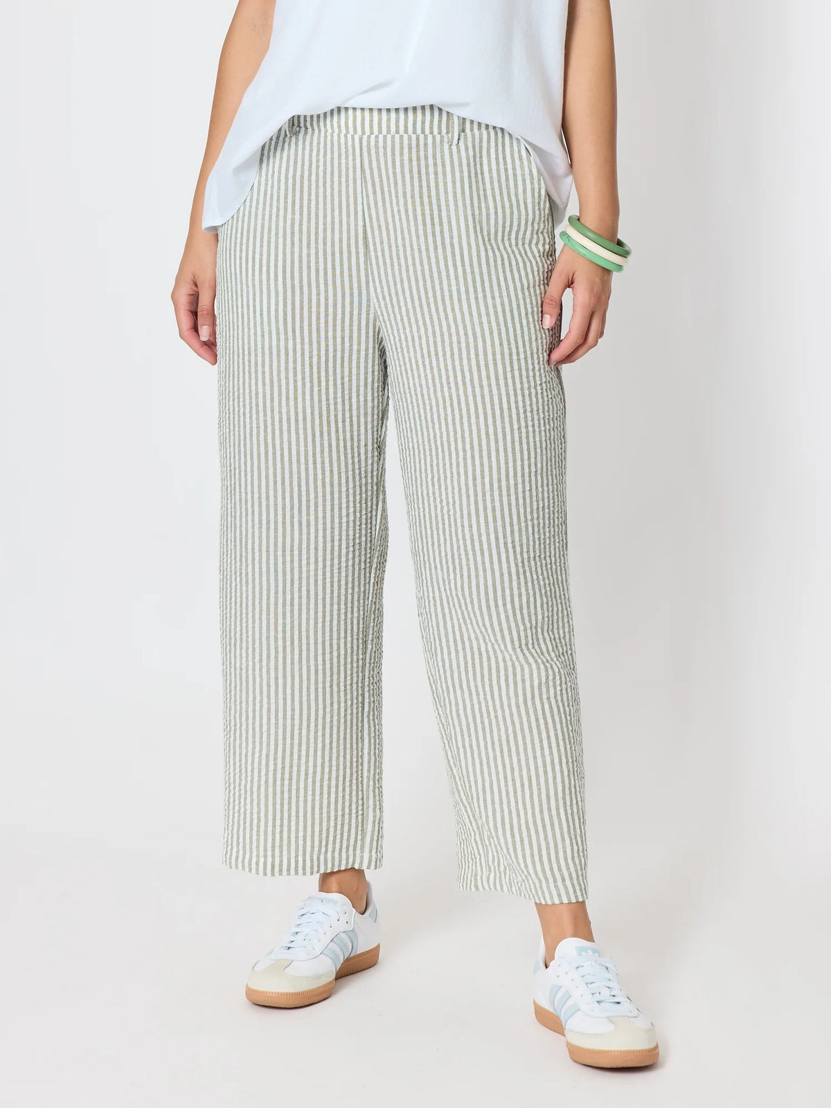 A woman wearing Threadz light green and white vertically striped cropped trousers, styled with a white top and white sneakers against a plain light gray studio background.