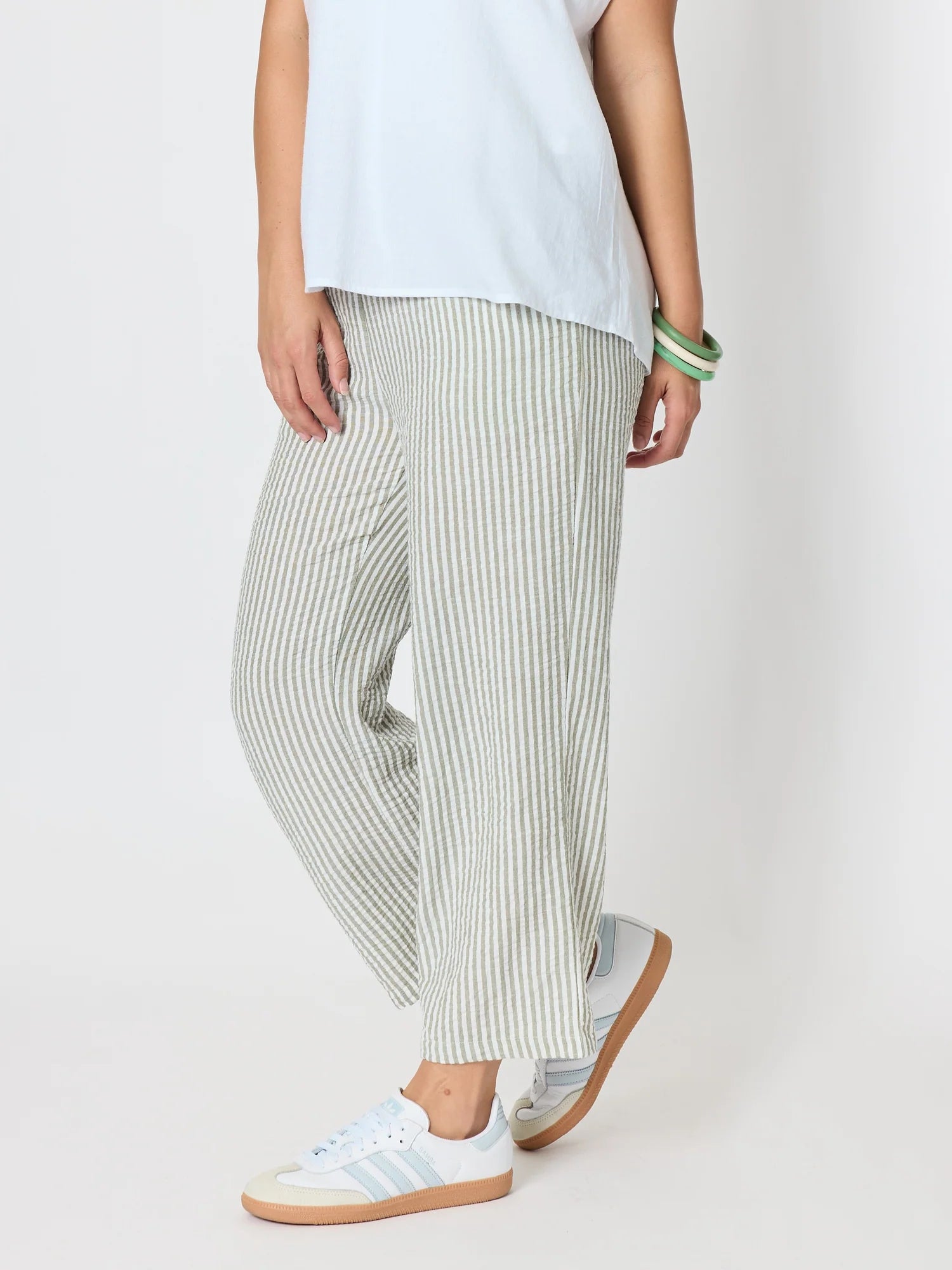 Image of A woman wearing Threadz loose, wide-leg striped pants in off-white and muted green, paired with a white top and white sneakers, photographed against a plain light background.
