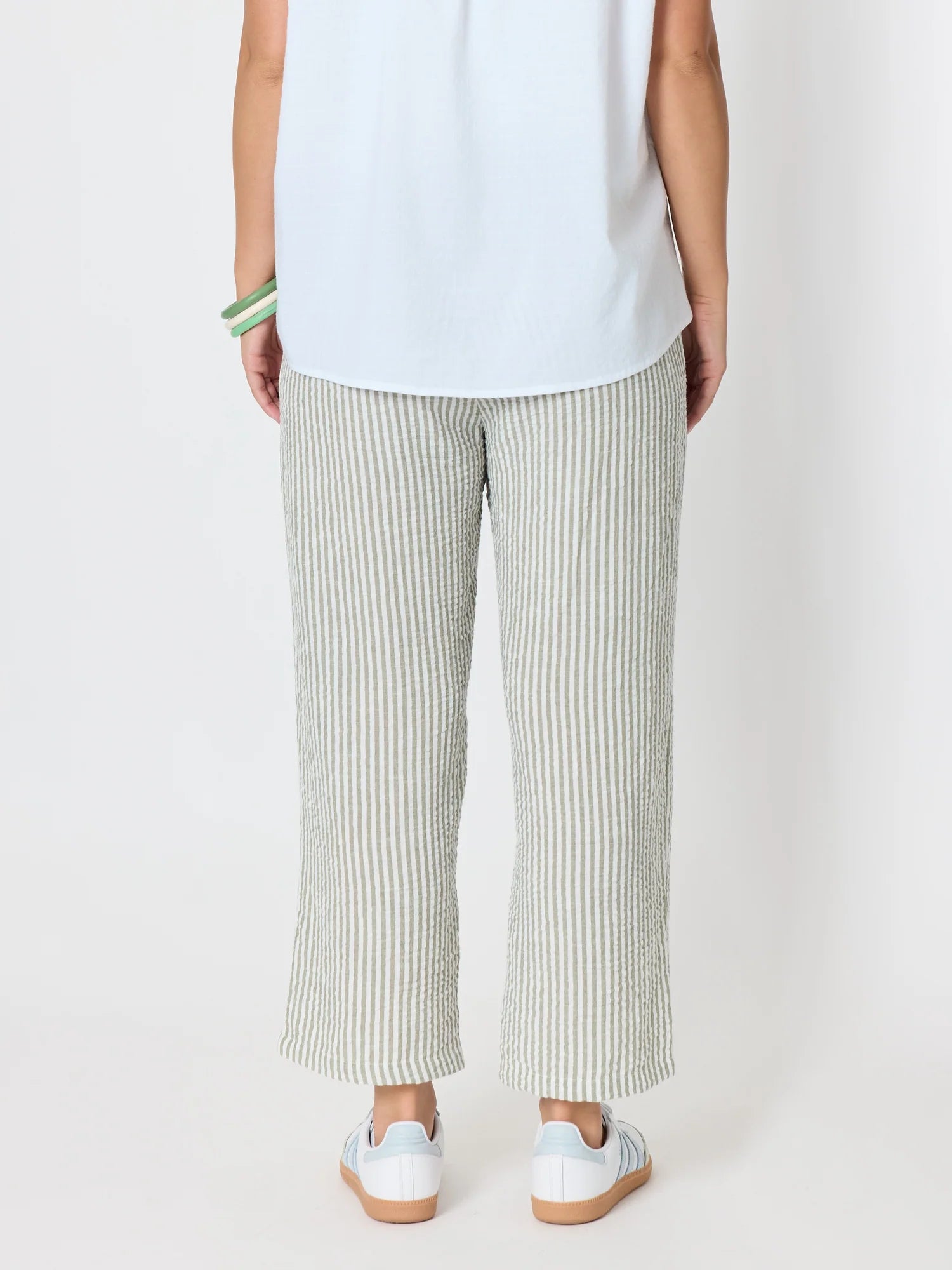 Image of A woman shown from the back wearing Threadz light green and white vertically striped wide-leg cropped pants, a white sleeveless top, and white sneakers with gum soles, against a plain light gray studio background.