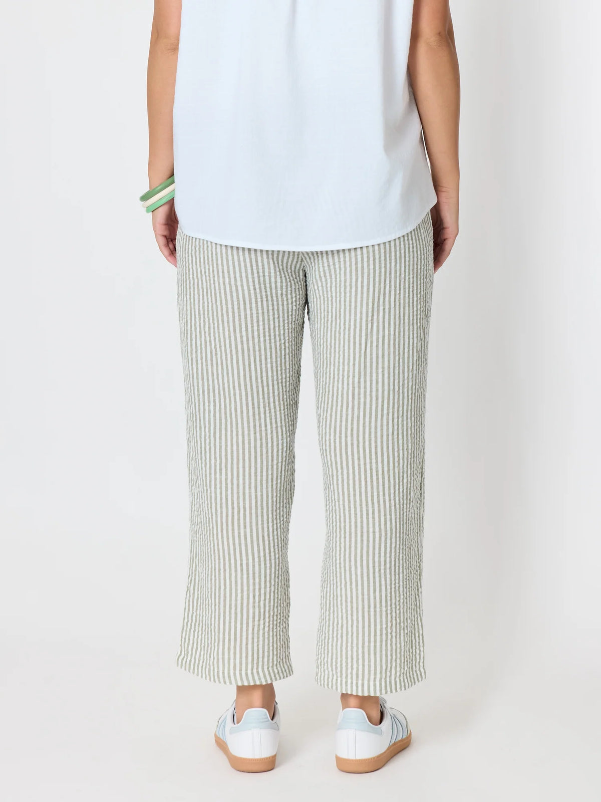 A woman shown from the back wearing Threadz light green and white vertically striped wide-leg cropped pants, a white sleeveless top, and white sneakers with gum soles, against a plain light gray studio background.